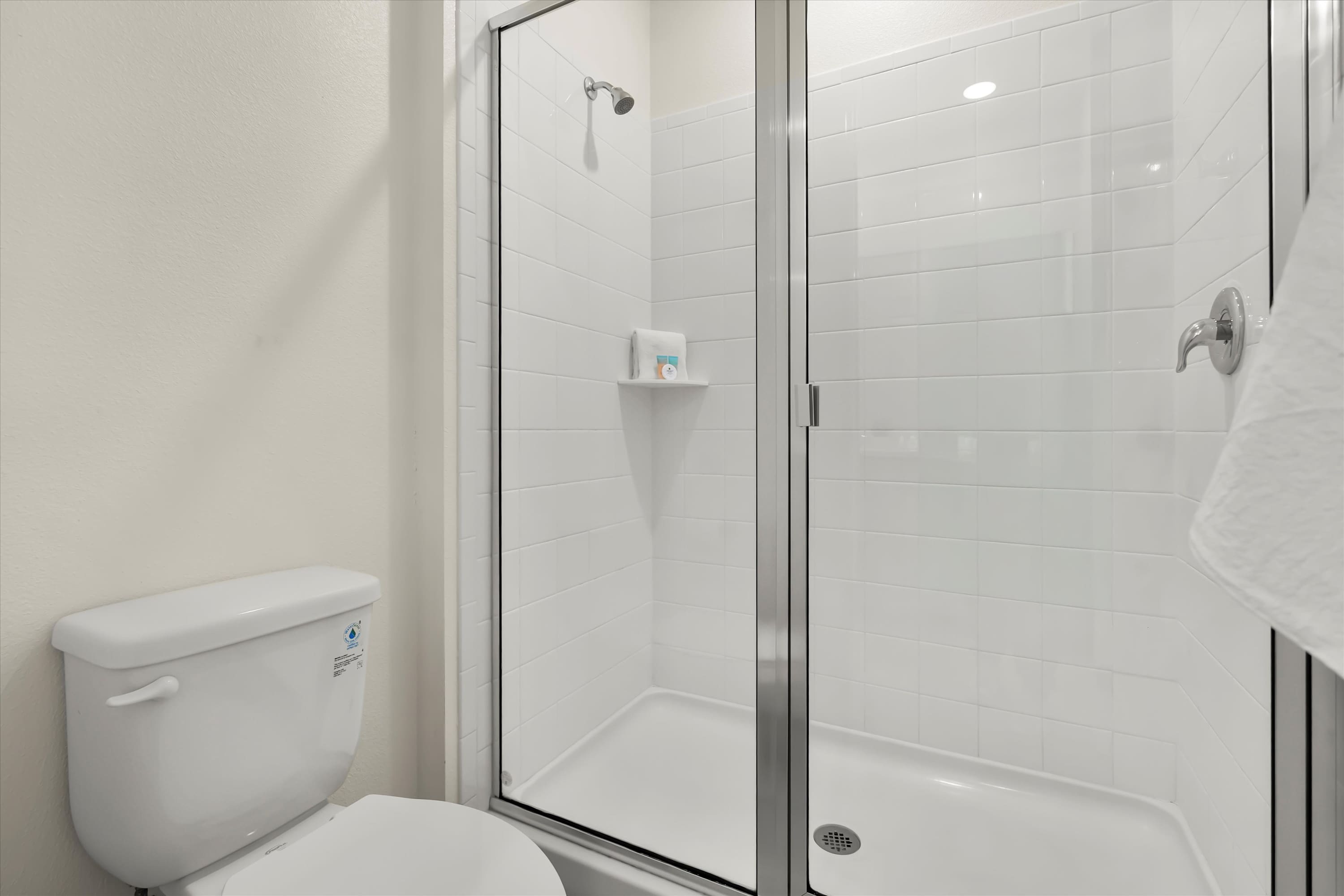 Close-up of a modern shower with white subway tiles and a clear glass enclosure, adjacent to a white toilet. A small shelf is visible inside the shower, and a white towel hangs on the wall.