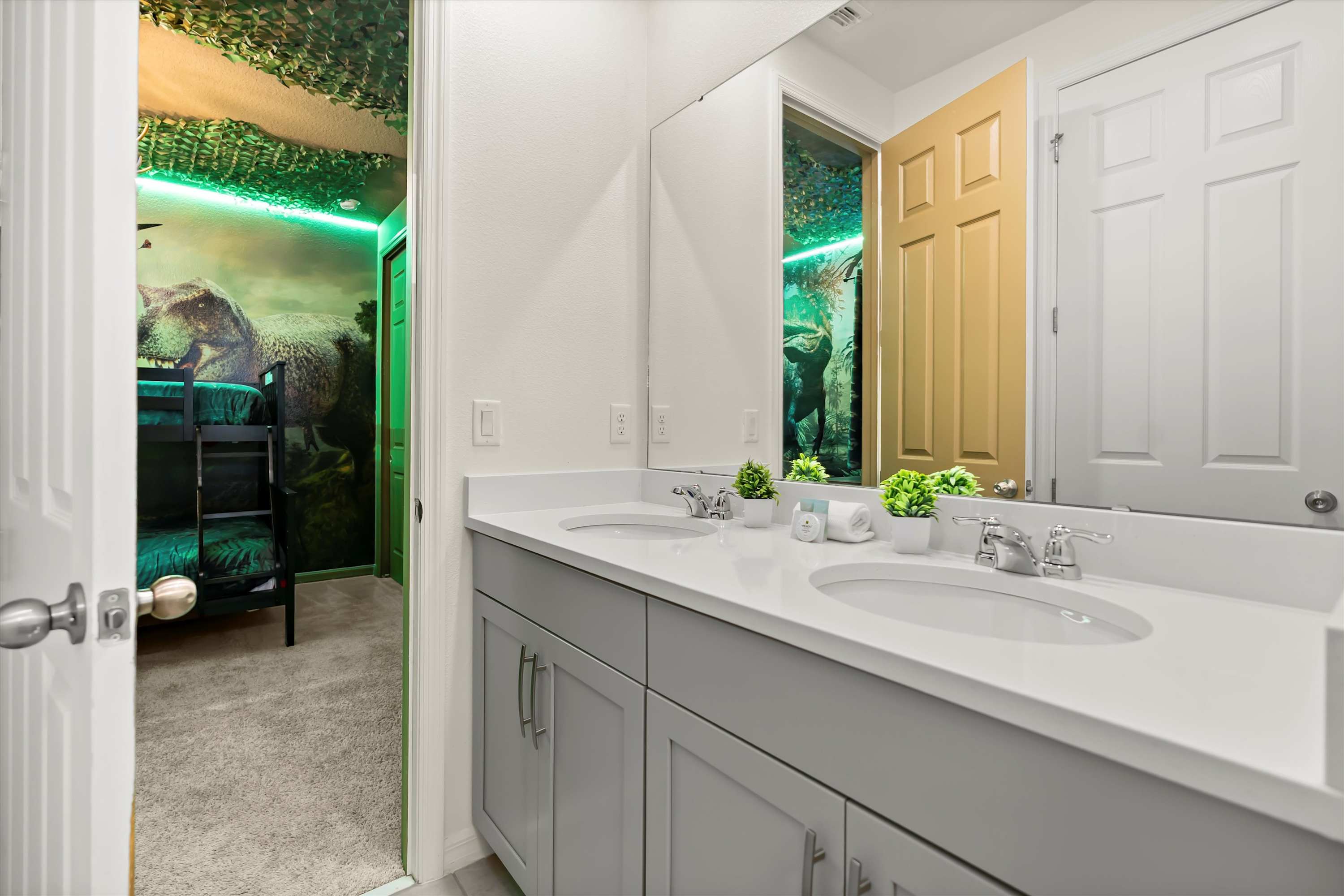 Modern bathroom with a double vanity featuring light gray cabinets and white countertops, reflecting a dinosaur-themed bunk bed room in the mirror and through the open doorway.