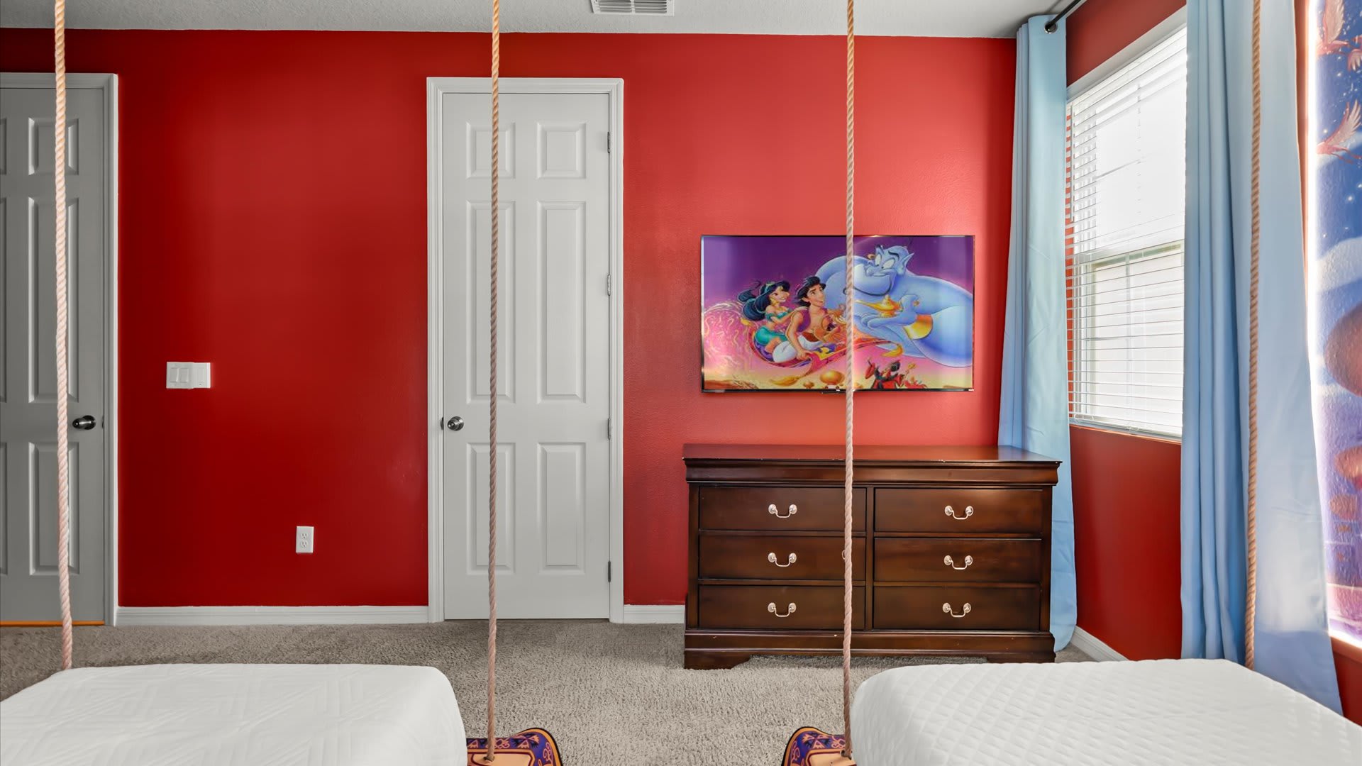 View of the Aladdin-themed bedroom showing a dark wood dresser with a mounted TV displaying an animated scene, red walls, and two white doors.