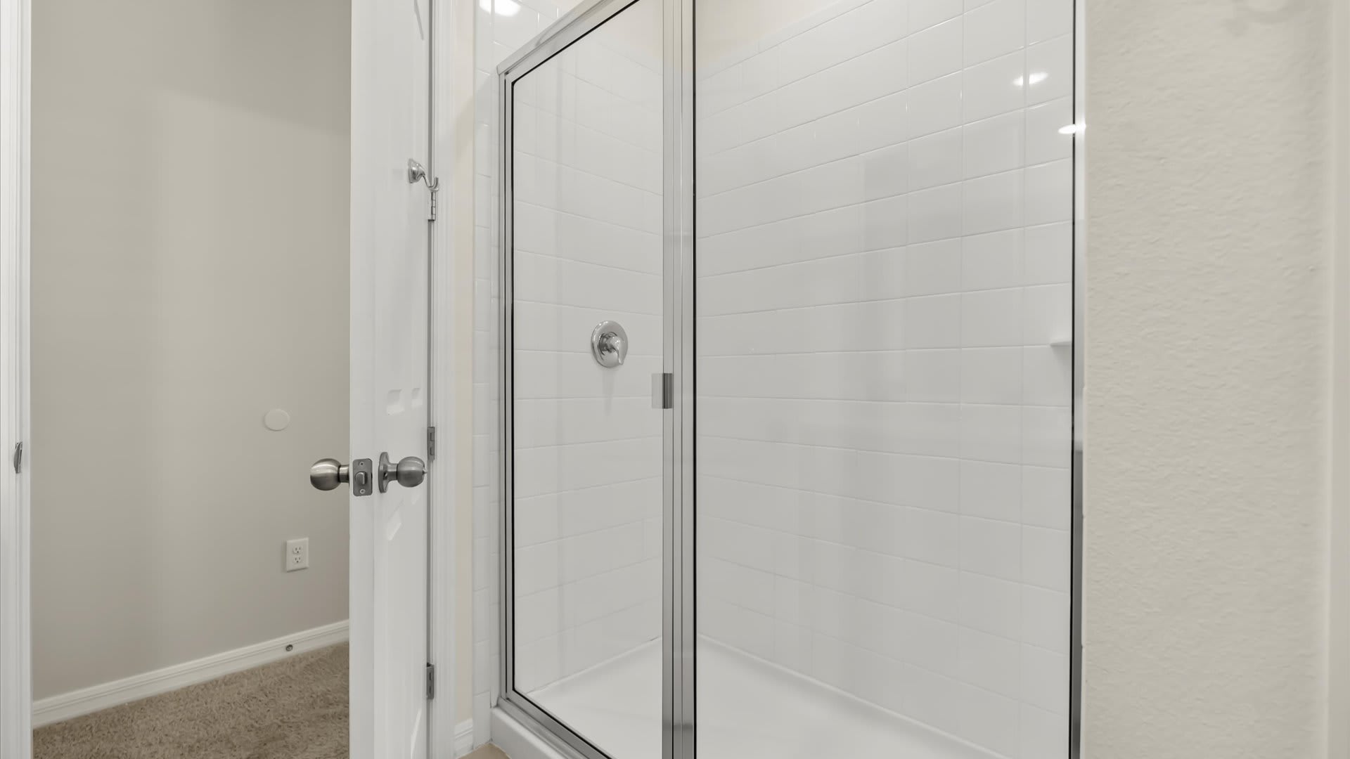 Close-up view of a modern shower with white subway tiles and a clear glass enclosure. A white shower pan and a wall-mounted shower head are visible, along with a small shelf for toiletries.
