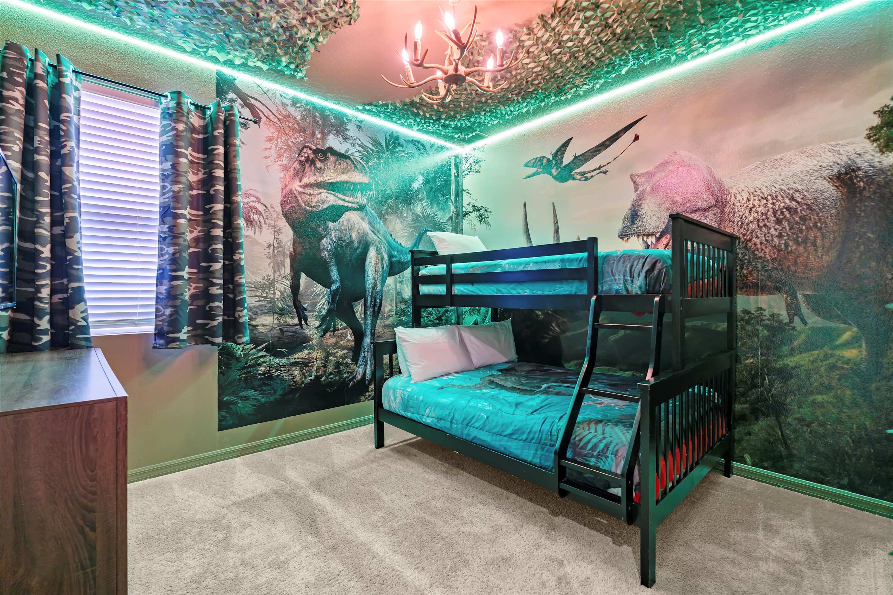 Dinosaur-themed bedroom featuring a black bunk bed with blue bedding, vibrant wall murals of dinosaurs, and green LED strip lighting along the ceiling with camouflage netting.