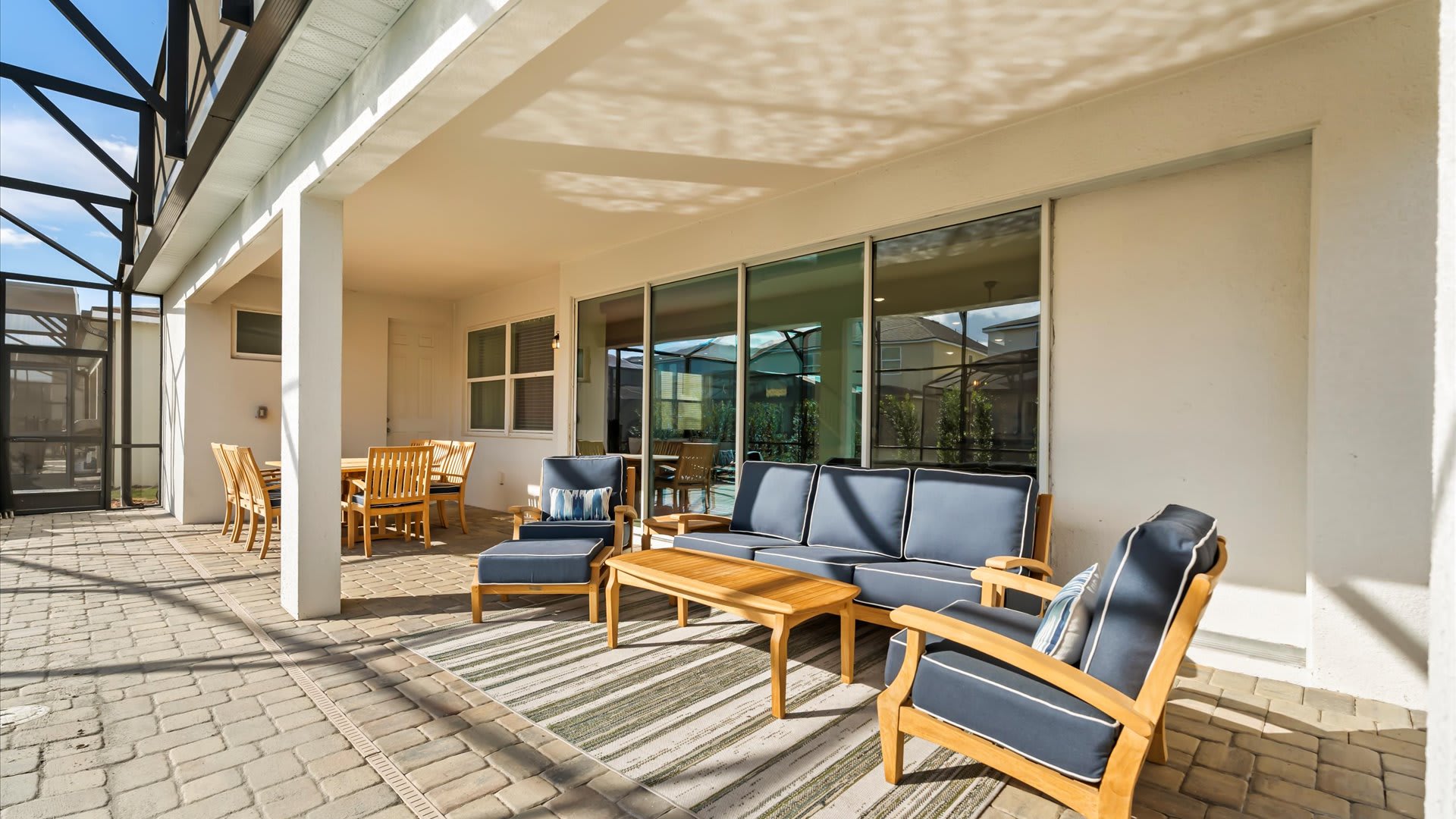 Covered outdoor patio featuring a comfortable seating arrangement with a blue sofa, two armchairs, and a coffee table, all made of wood with blue cushions, situated on a striped rug next to a screened pool area.