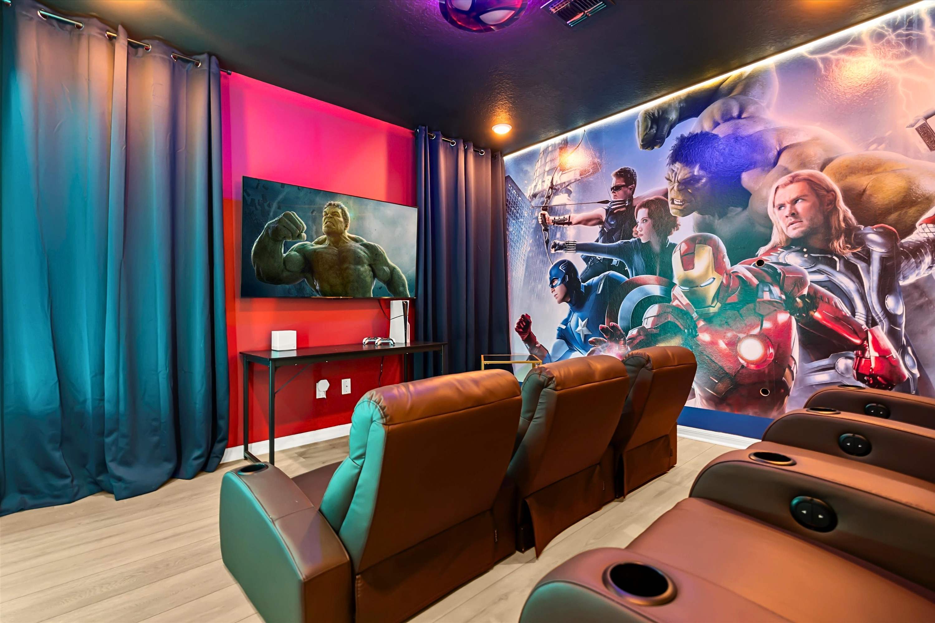 A themed media room features rows of brown leather recliners facing a large wall-mounted TV displaying a Hulk image, with a vibrant Marvel superhero mural covering the adjacent wall. Dark curtains frame the TV area, and the ceiling is dark with colorful accent lighting.