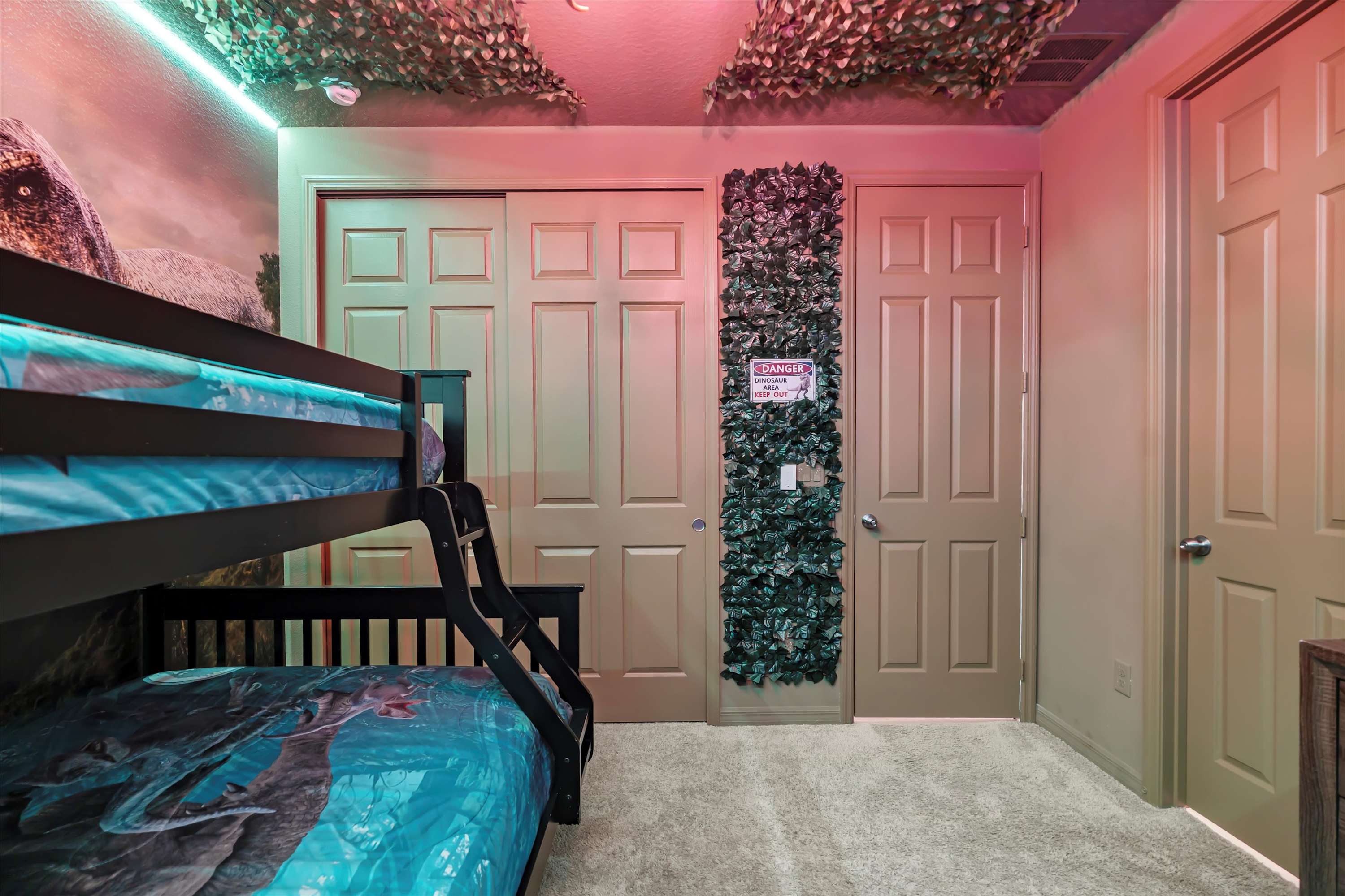 View of the dinosaur-themed bedroom showing a black bunk bed with blue bedding, closet doors, and a "DANGER DINOSAUR CROSSING" sign on a vine-covered wall panel. Green LED lighting illuminates the camouflage ceiling.