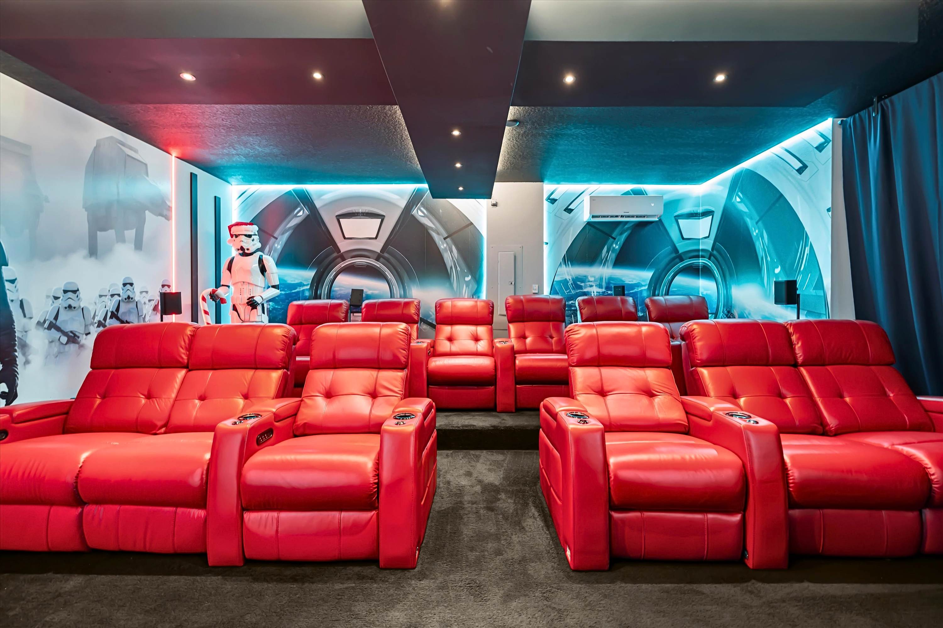 A Star Wars-themed media room features two rows of plush red leather recliners on dark carpet, facing a wall with a large mural depicting Stormtroopers and a futuristic tunnel. The dark ceiling has recessed lighting, and blue accent lighting illuminates the walls.