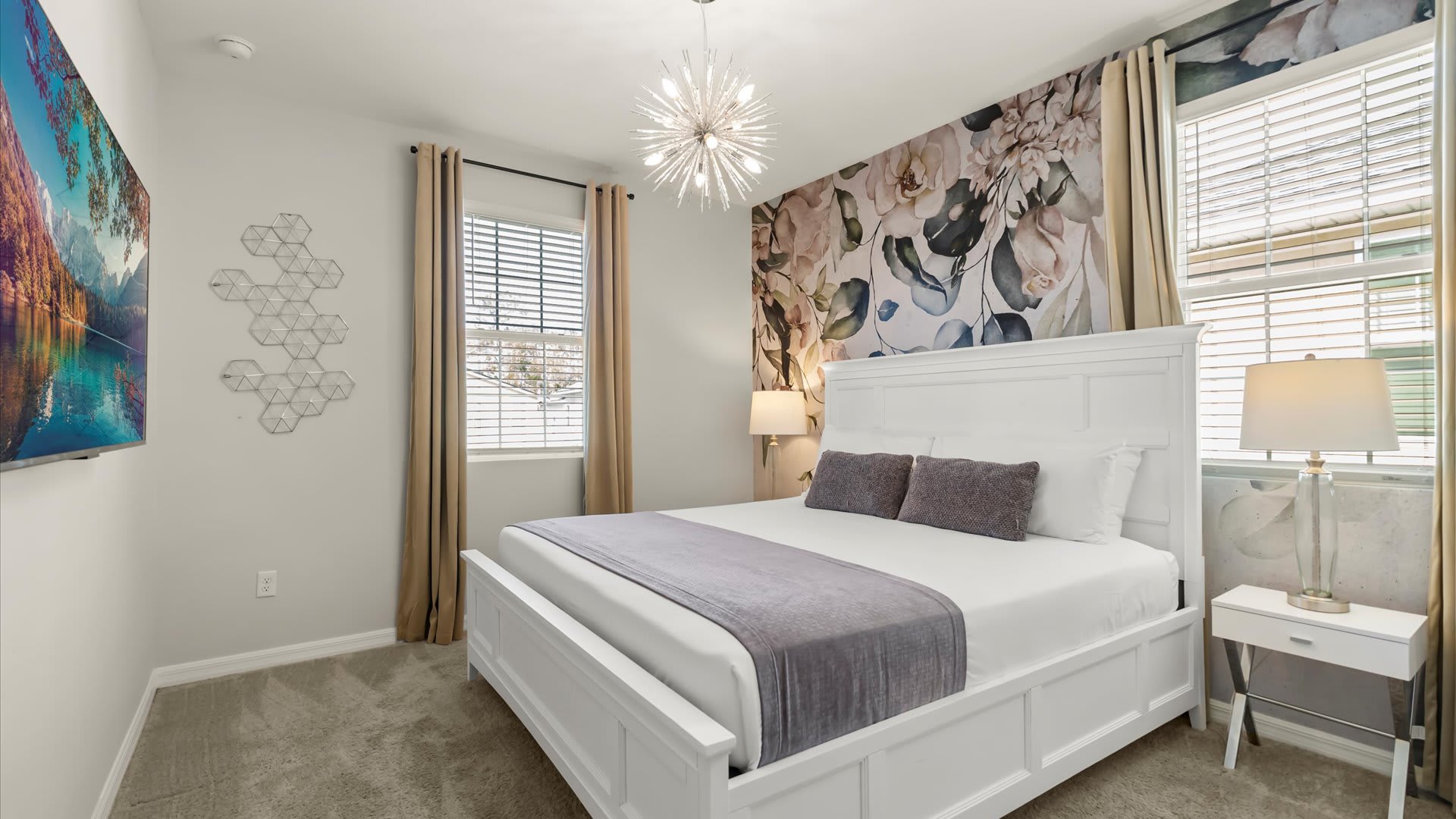Elegant bedroom featuring a white bed frame, a floral accent wall behind the headboard, and a unique sputnik-style chandelier. Two windows with blinds are draped with beige curtains, and a wall-mounted TV is visible on the opposite wall.