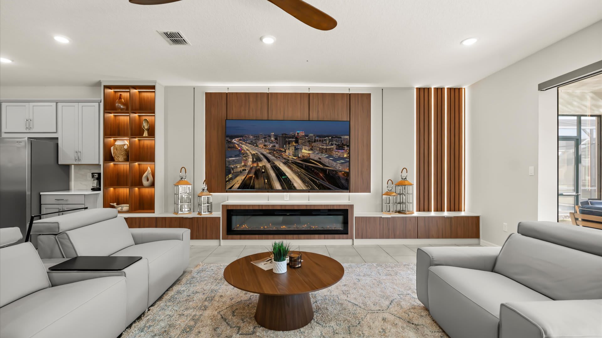 A modern living room features two light gray sofas facing a large wall-mounted TV above an electric fireplace, set within a stylish wood-paneled media console with integrated shelving. A round wooden coffee table sits on a patterned rug, illuminated by recessed lighting and a ceiling fan.