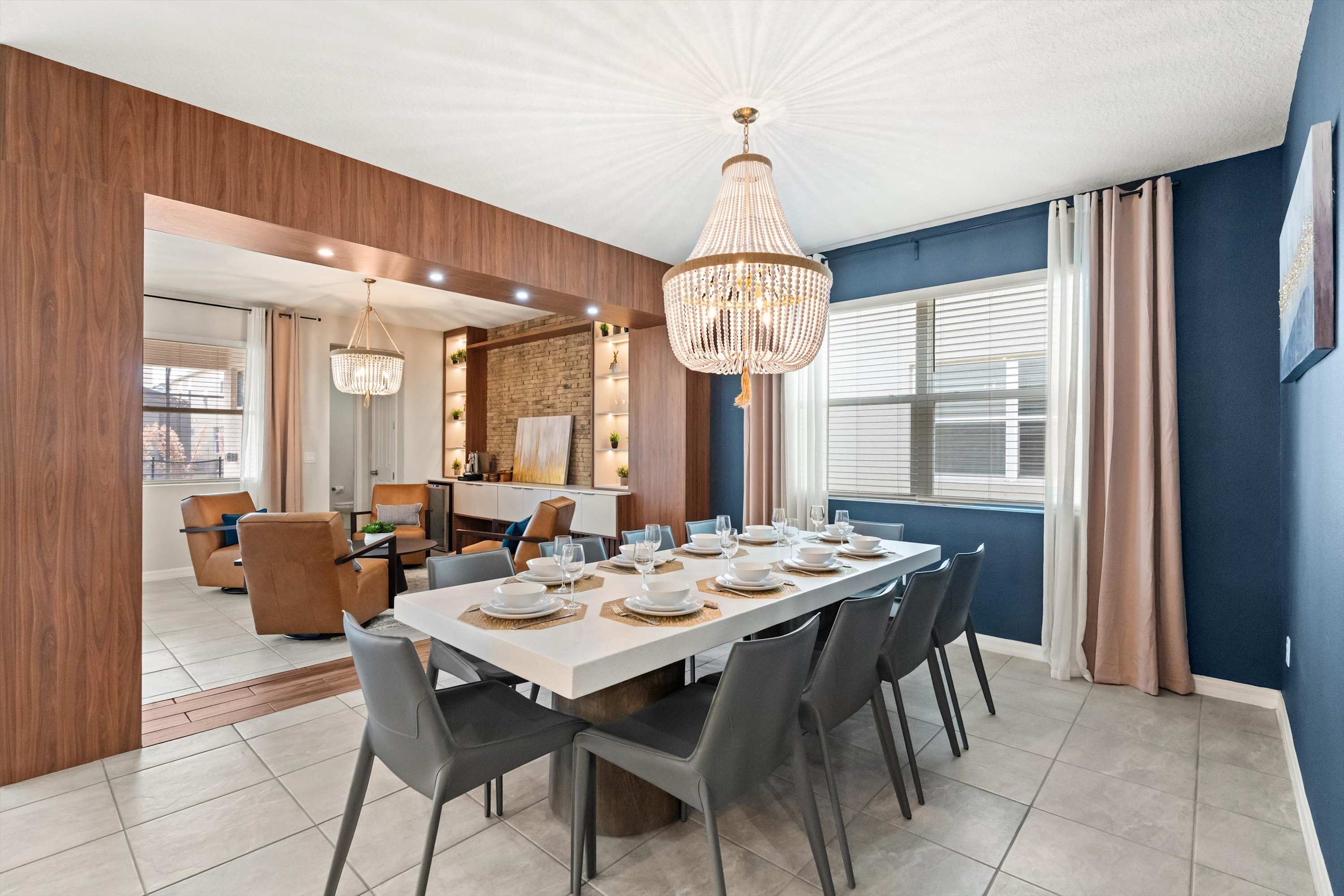 The dining room features a long white table with eight dark gray chairs, set beneath a large crystal chandelier. A dark blue accent wall is visible, and a reflection in the mirror shows the adjacent lounge area with leather chairs and a brick wall.