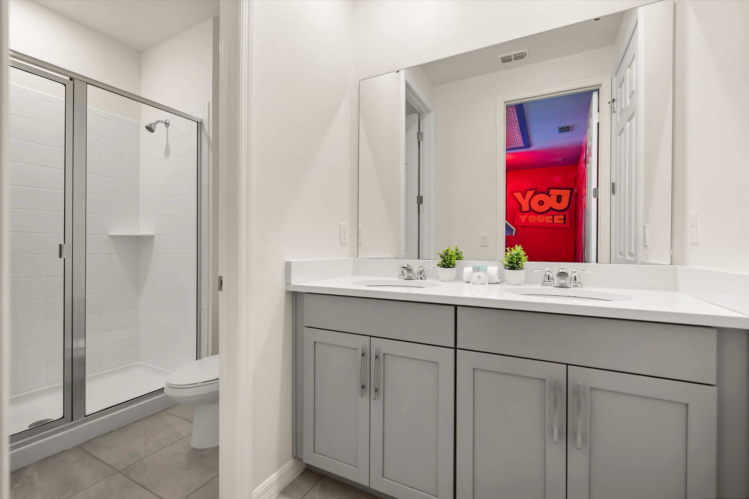 Modern bathroom with a double vanity featuring light gray cabinets and white countertops, a walk-in shower with glass door, and a reflection of the Toy Story themed room in the mirror.