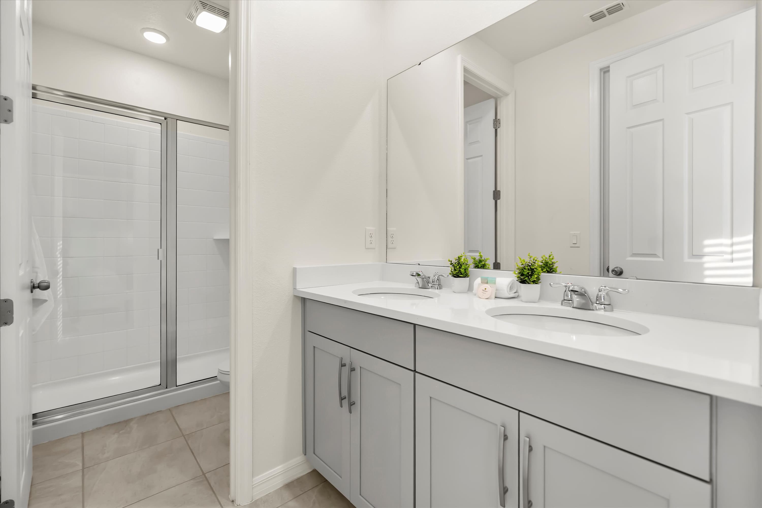 Modern bathroom featuring a double vanity with light gray cabinets, white countertops, and two rectangular sinks. A large mirror spans the width of the vanity, reflecting a white door and a shower with a glass enclosure.