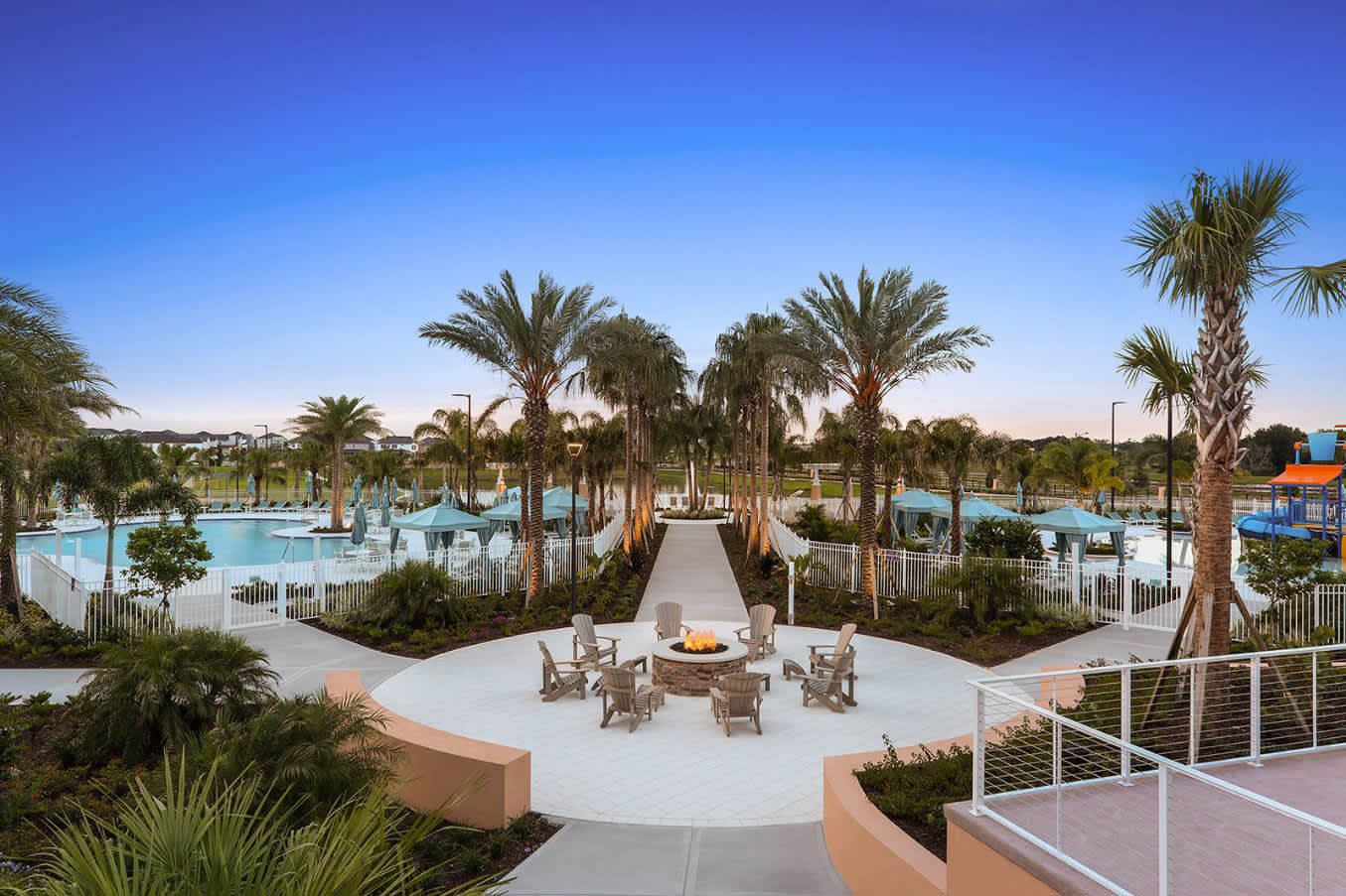 An expansive resort pool area is visible at dusk, featuring a large communal pool with cabanas, surrounded by numerous palm trees, and a central circular fire pit with Adirondack chairs.