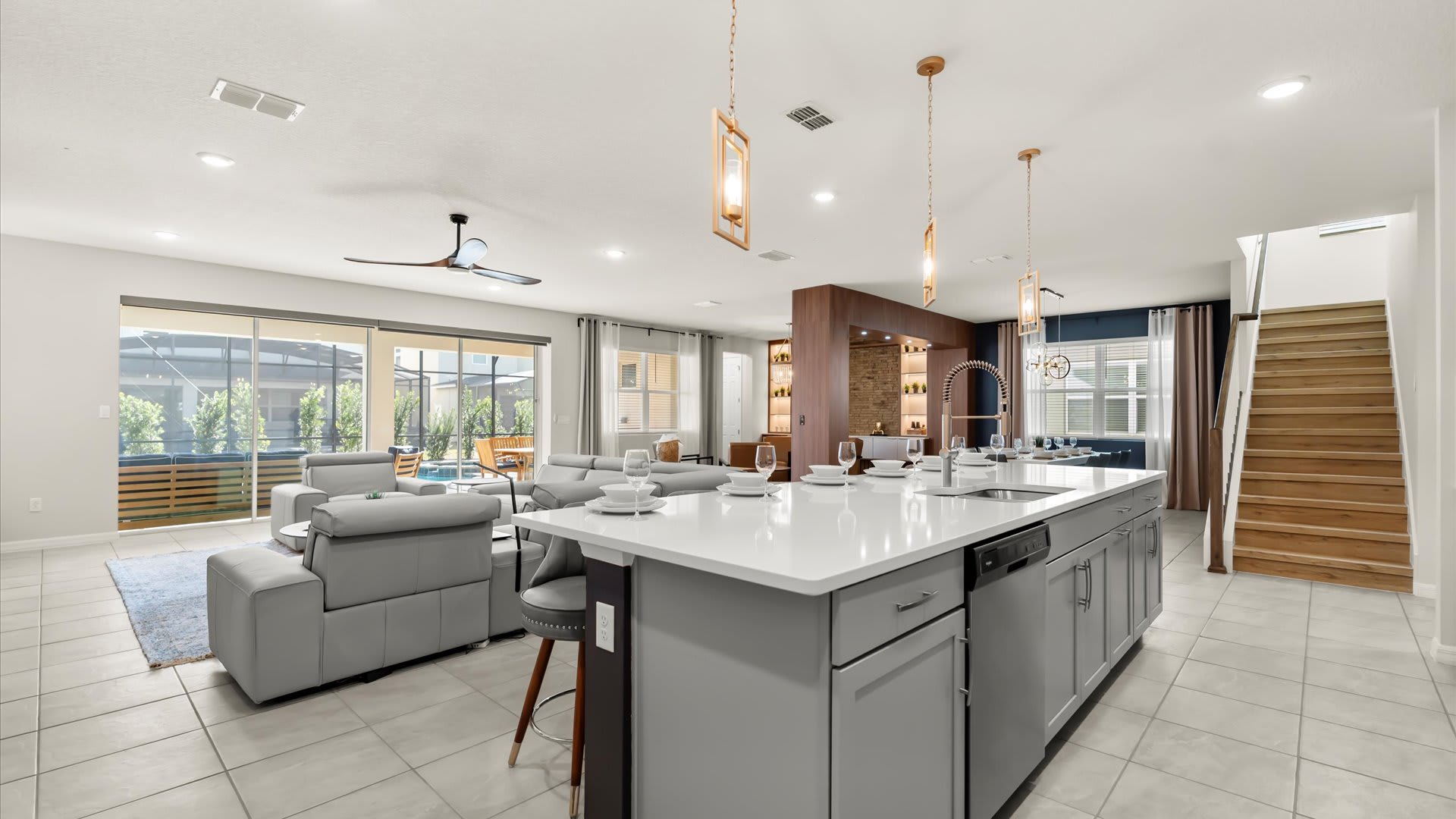 An open-concept kitchen features a large island with bar seating and pendant lights, overlooking a spacious living room with a view of the outdoor pool area through sliding glass doors. A wooden staircase is visible on the right, connecting the levels of the home.