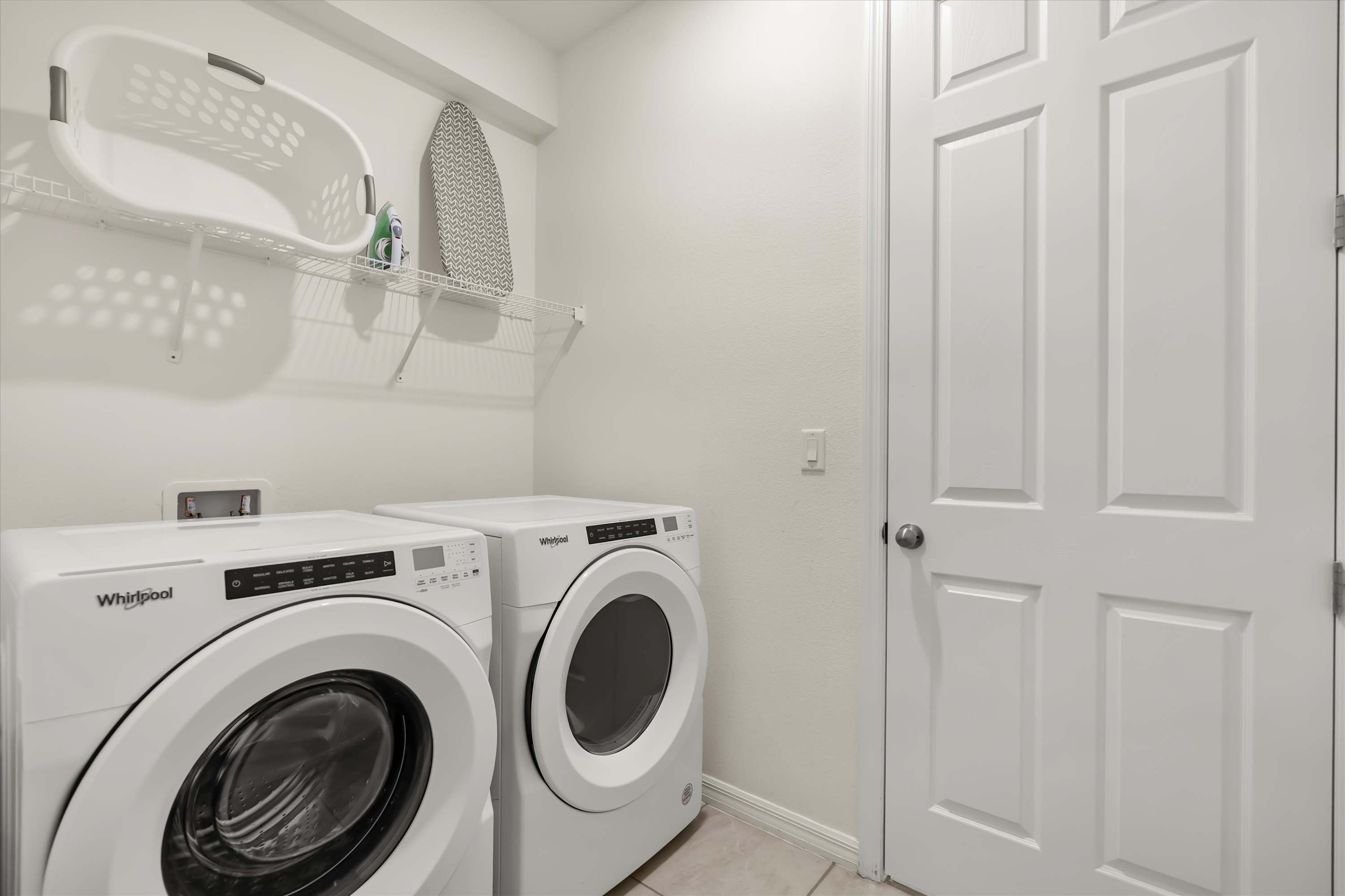 Bright laundry room equipped with a white Whirlpool washer and dryer, a wire shelf with a laundry basket and ironing board, and light-colored walls.