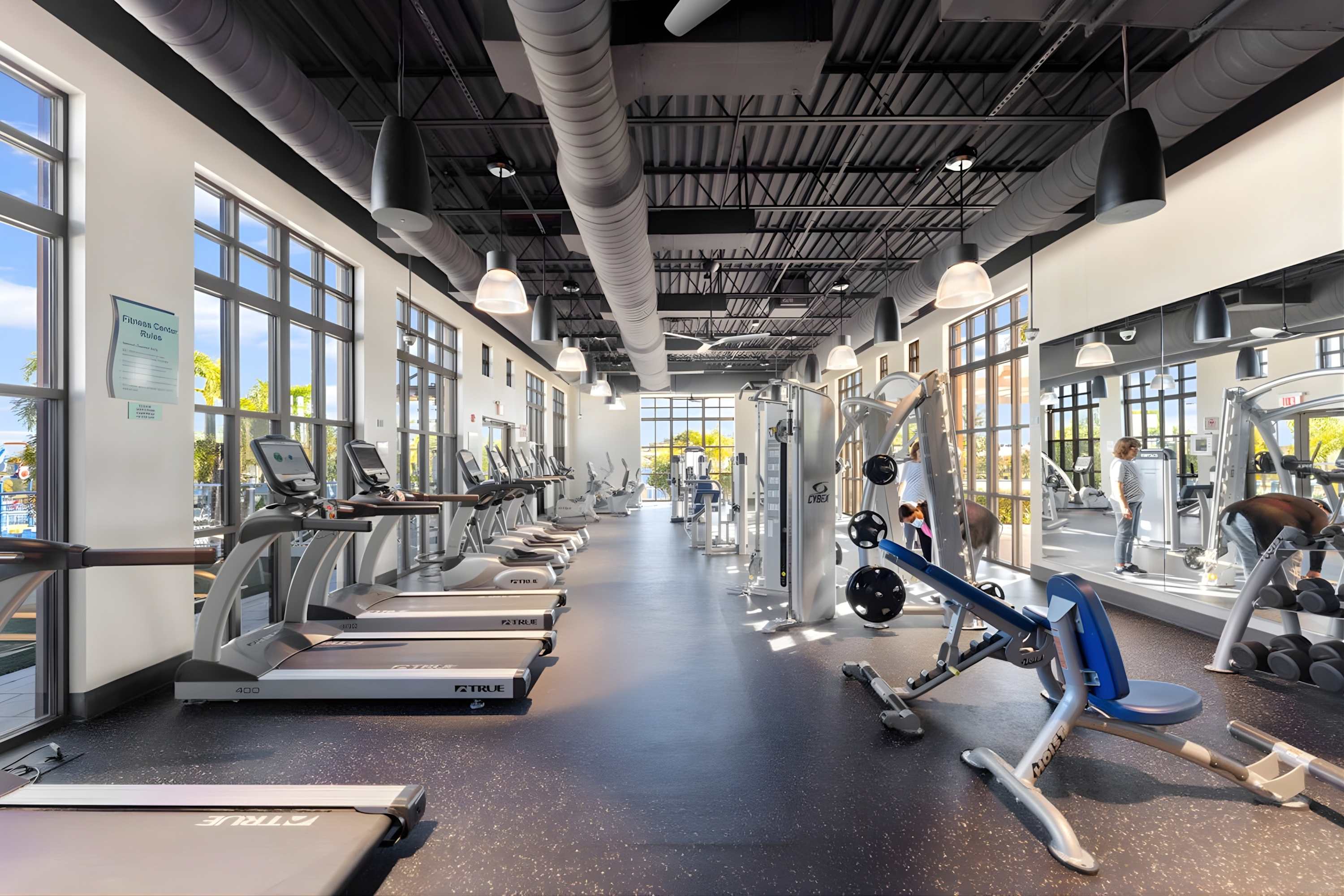 A spacious, modern fitness center features rows of treadmills and various weight machines, with large windows offering natural light and views of the outdoor pool area. The industrial-style ceiling has exposed ducts.