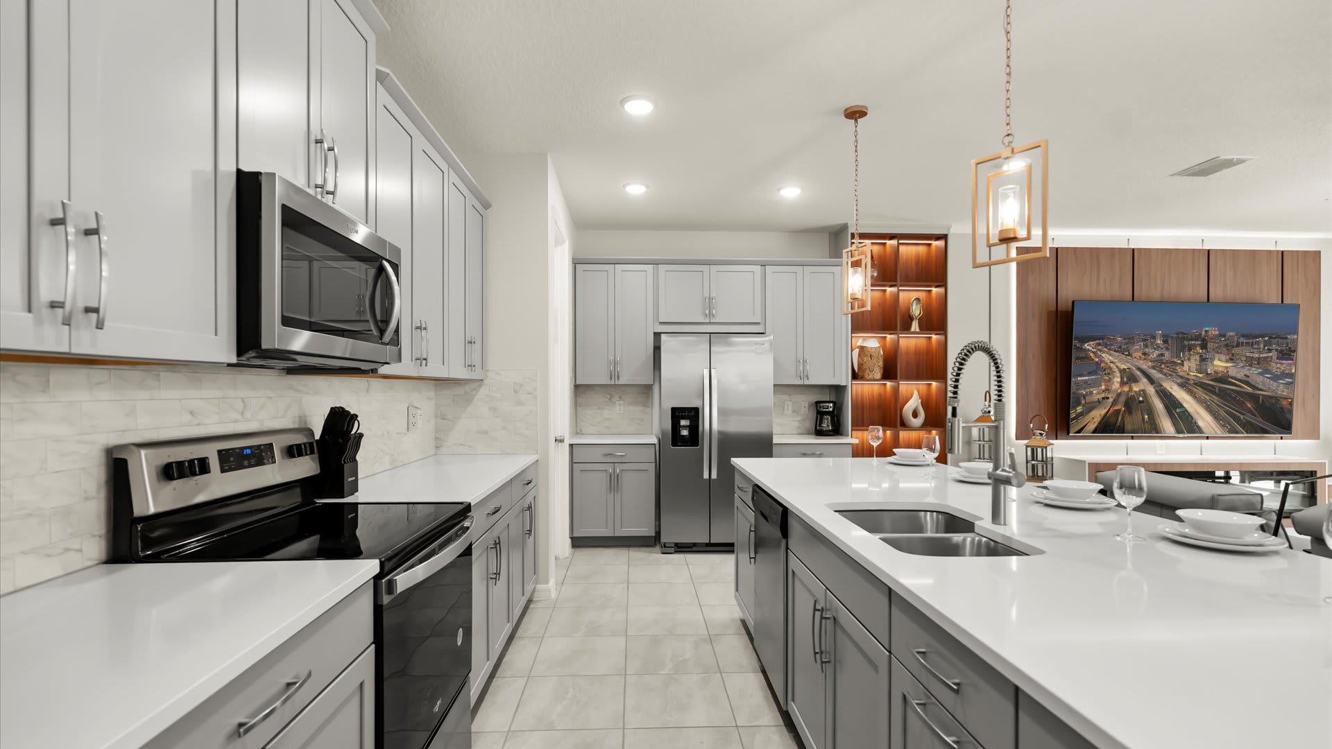This view of the kitchen highlights light gray shaker cabinets, white countertops, and stainless steel appliances, including a microwave, stove, and refrigerator. A long kitchen island with a sink and seating extends into the foreground, with the living room visible beyond.