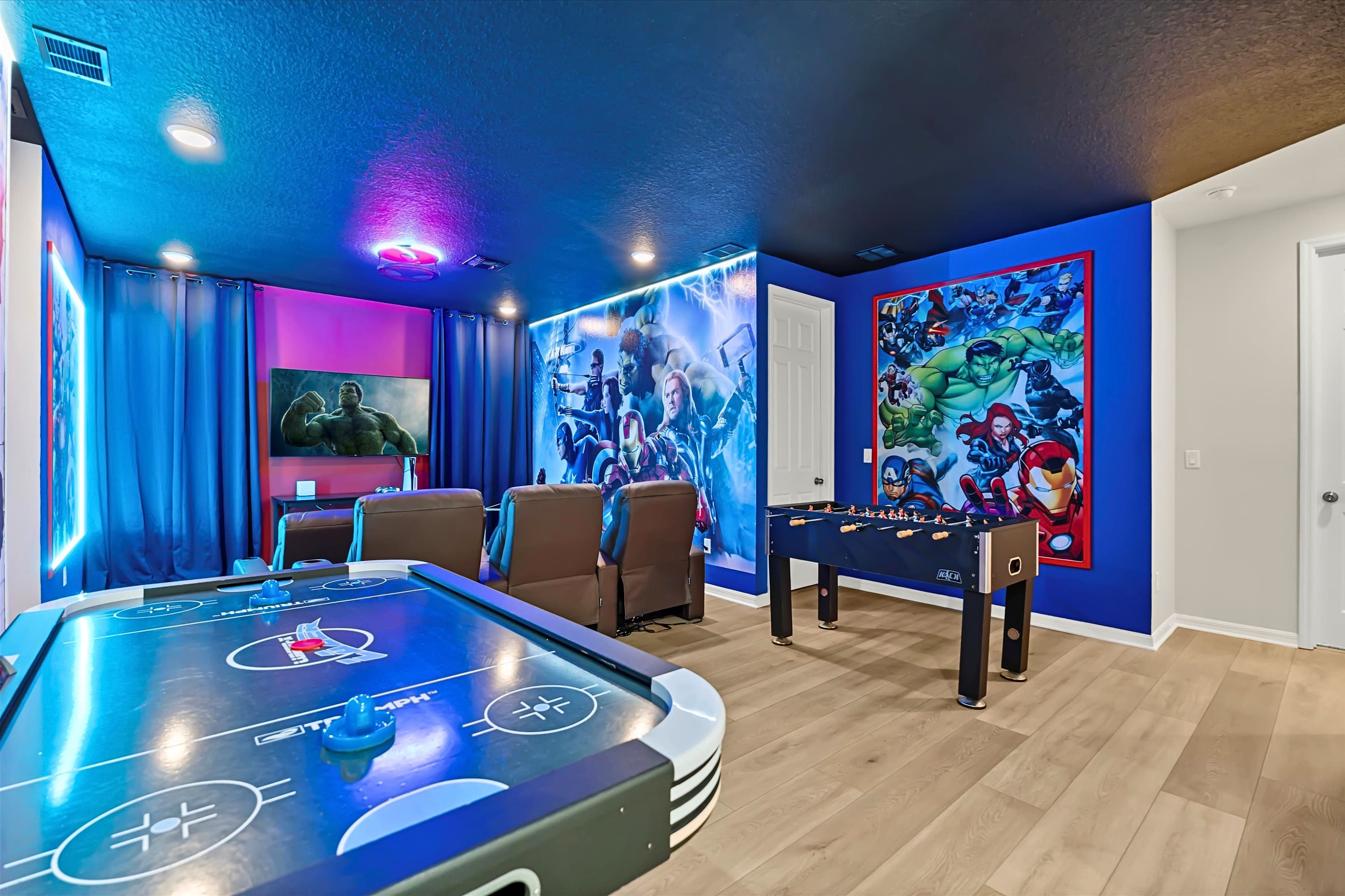Vibrant game room featuring an air hockey table, a foosball table, and a TV, all set against walls adorned with colorful Marvel superhero murals and illuminated by blue and purple LED lighting.