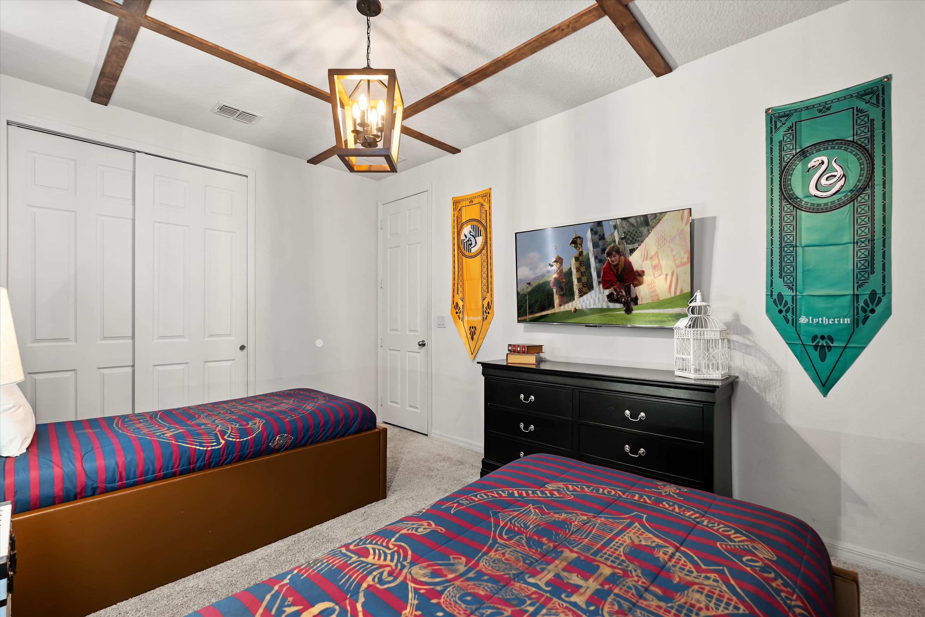 Harry Potter-themed bedroom with a black dresser, mounted TV, and two white closet doors, flanked by house banners and a window with blinds.