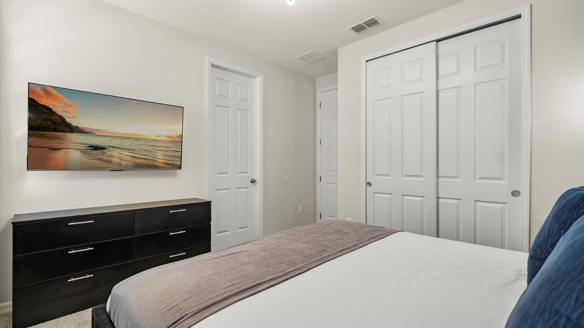 View of bedroom-3 showing a wall-mounted TV displaying a beach scene, a dark dresser, and white bi-fold closet doors. The room has light gray walls and a dark upholstered bed.