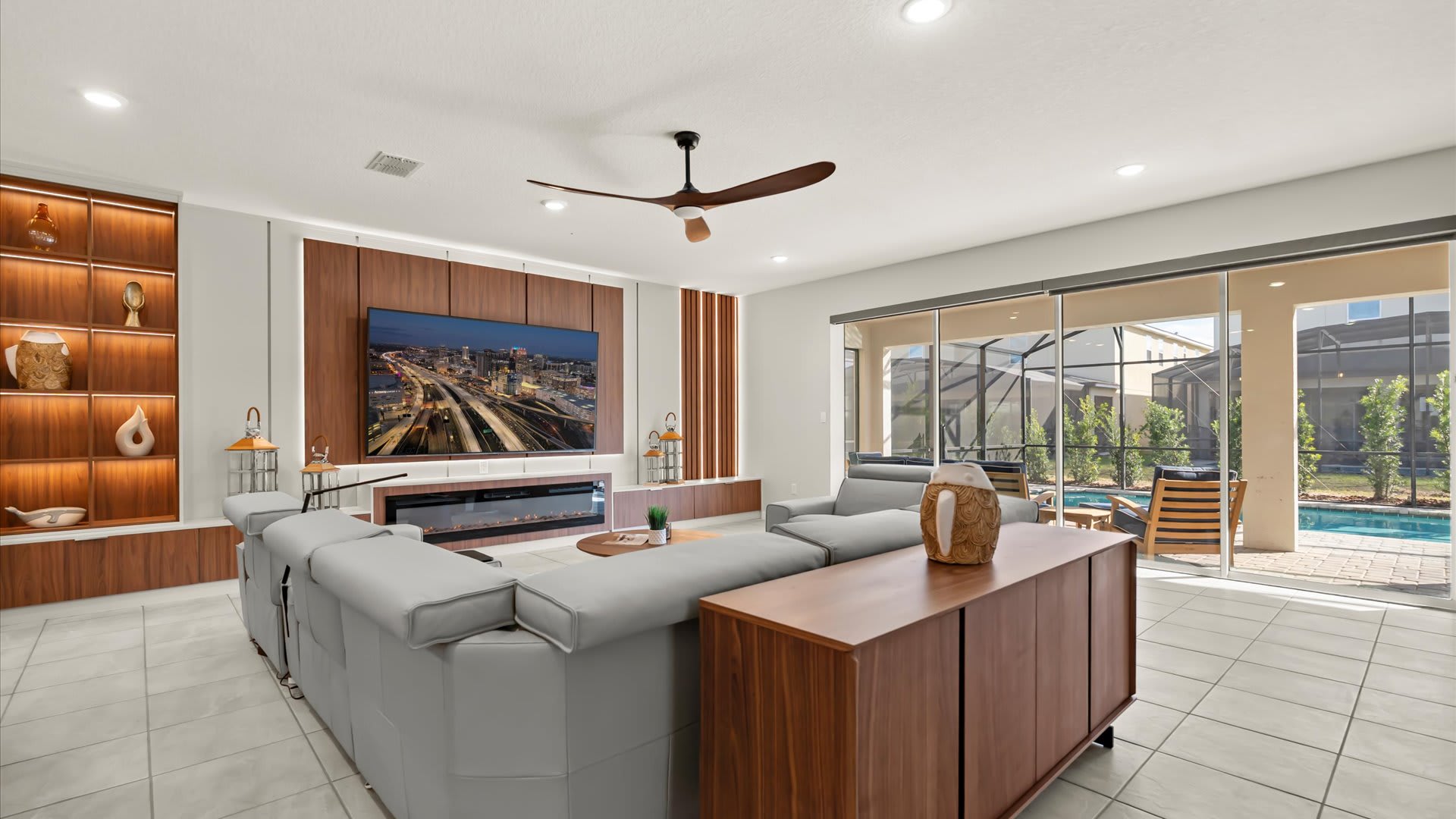This wide-angle view of the living room shows light gray sofas, a round wooden coffee table on a patterned rug, and a large TV mounted on a wood-paneled wall above an electric fireplace. Sliding glass doors open to an outdoor patio with a pool, visible in the background.