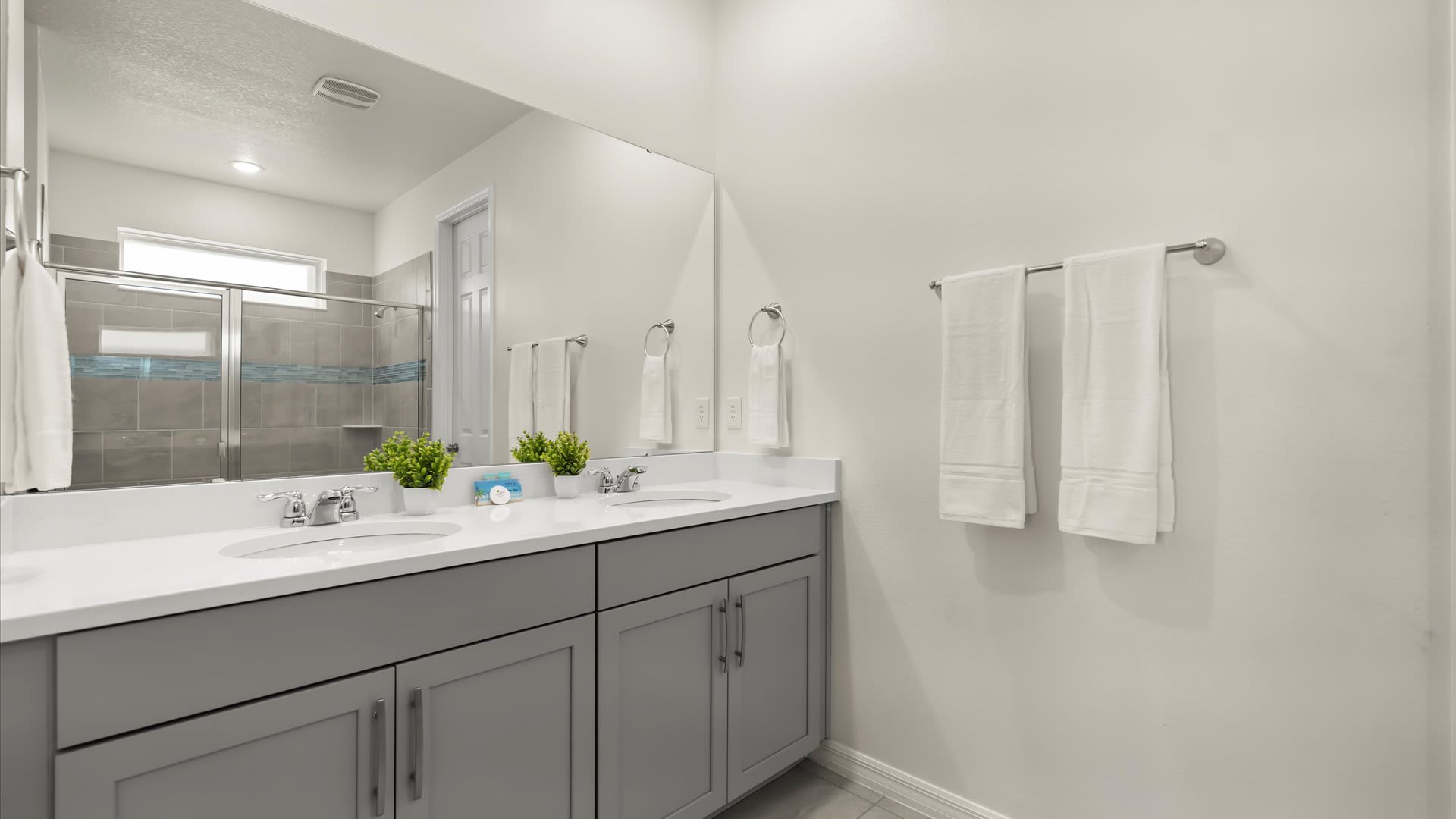 A bright bathroom features a double vanity with grey cabinets, white countertops, and two undermount sinks, beneath a large mirror. White towels hang on a rack, and a walk-in shower is visible in the background.