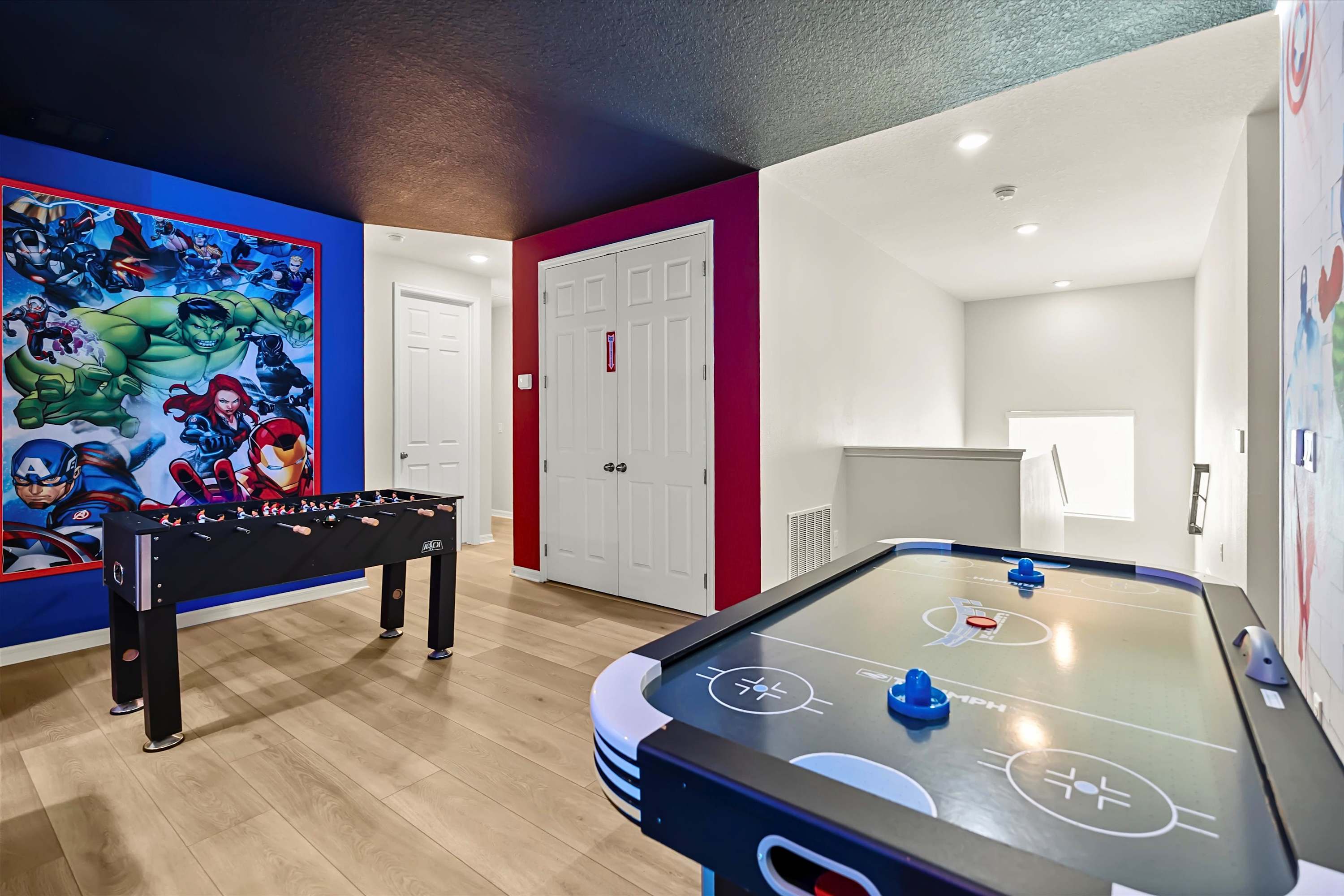 A vibrant game room features a foosball table and an air hockey table on light wood-look flooring, set against walls with a large Marvel superhero mural and a bold red accent wall. The ceiling is dark, and a white door is visible in the background.