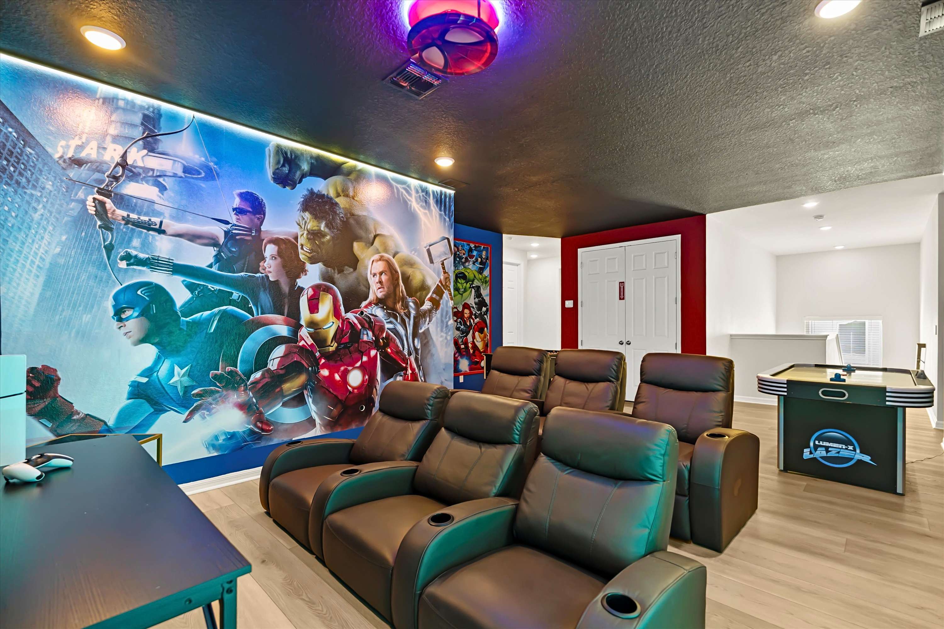 A spacious media room features multiple rows of brown leather recliners facing a large Marvel superhero mural, with an air hockey table visible in the background. The room has light wood-look flooring, a dark ceiling with recessed lighting, and a red accent wall.