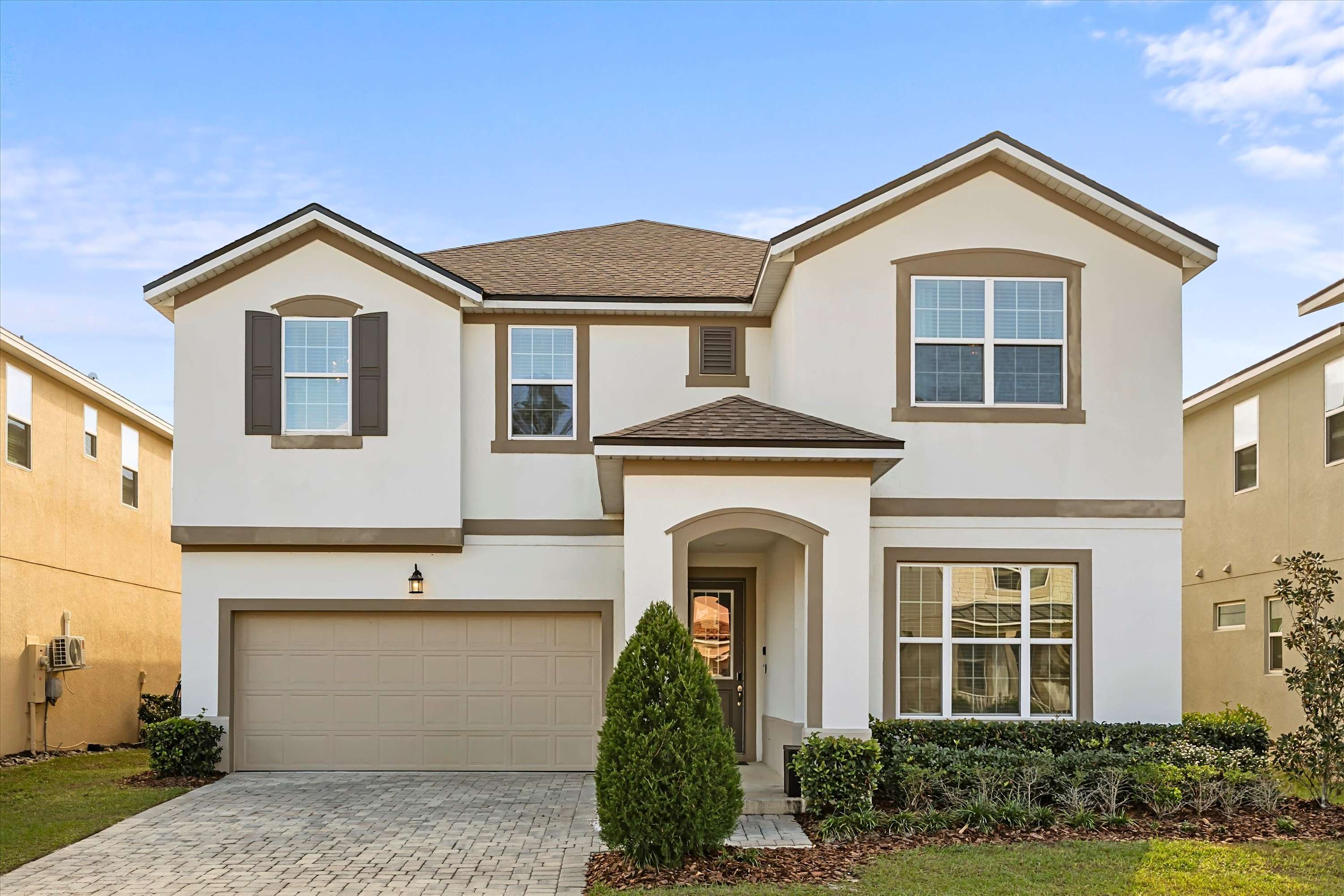 Two-story modern house with a light stucco exterior, brown trim, a two-car garage, and a covered entryway, set against a clear blue sky. A paved driveway and landscaped front yard are visible.