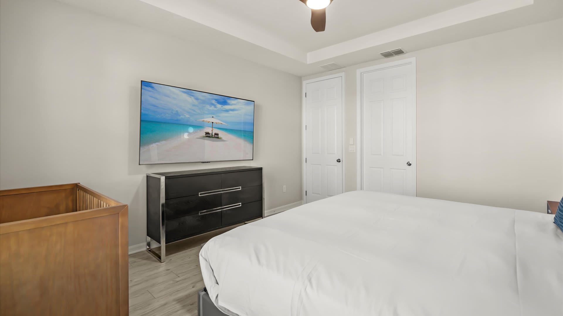 This bright bedroom features a large wall-mounted TV above a black dresser with silver accents, opposite a king-sized bed. A wooden crib is visible in the foreground, and the room has light wood-look flooring and a ceiling fan.