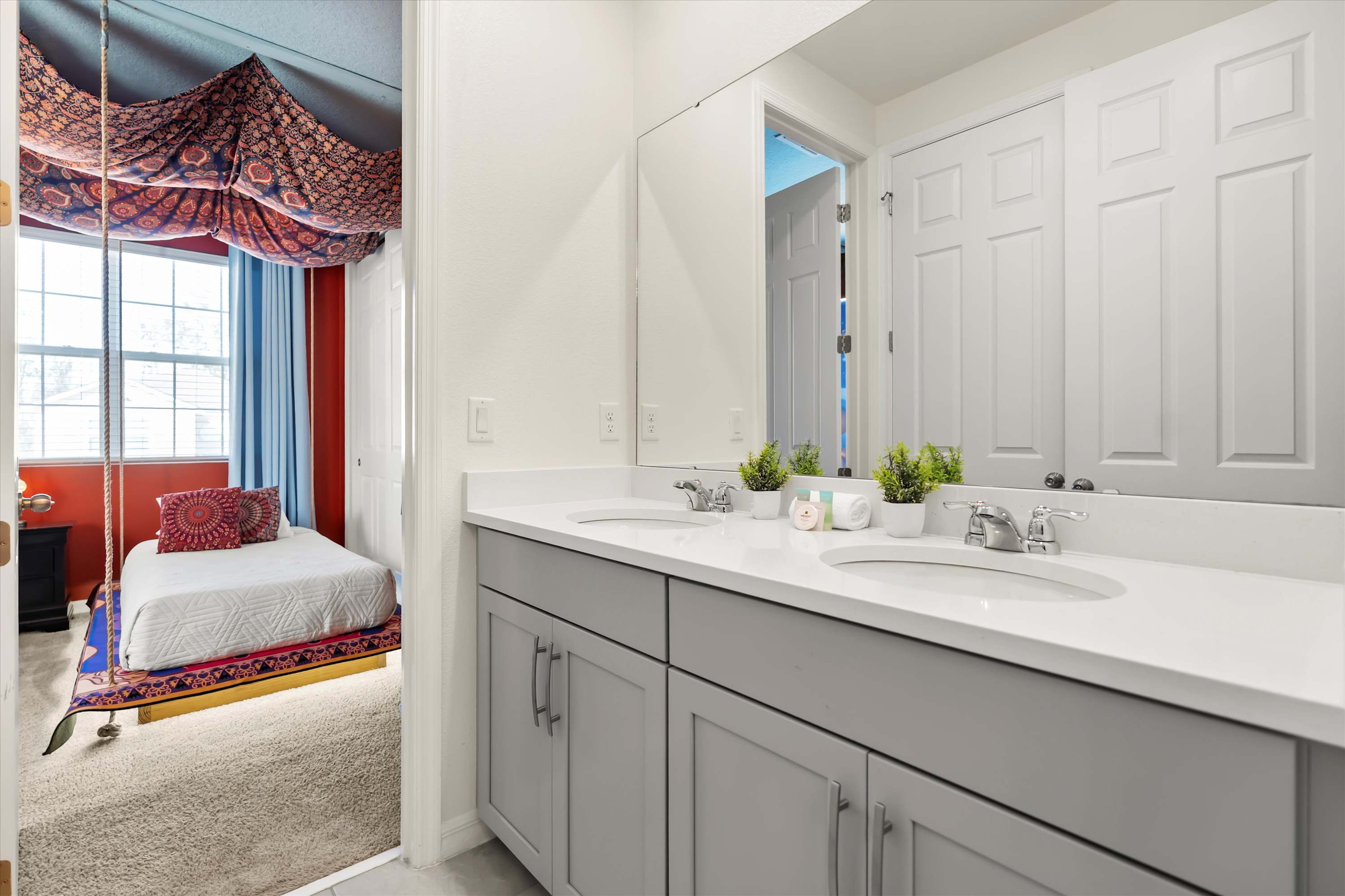 Modern bathroom with a light gray double vanity, white quartz countertop, and a large mirror reflecting a themed bedroom with a hanging bed and red walls.