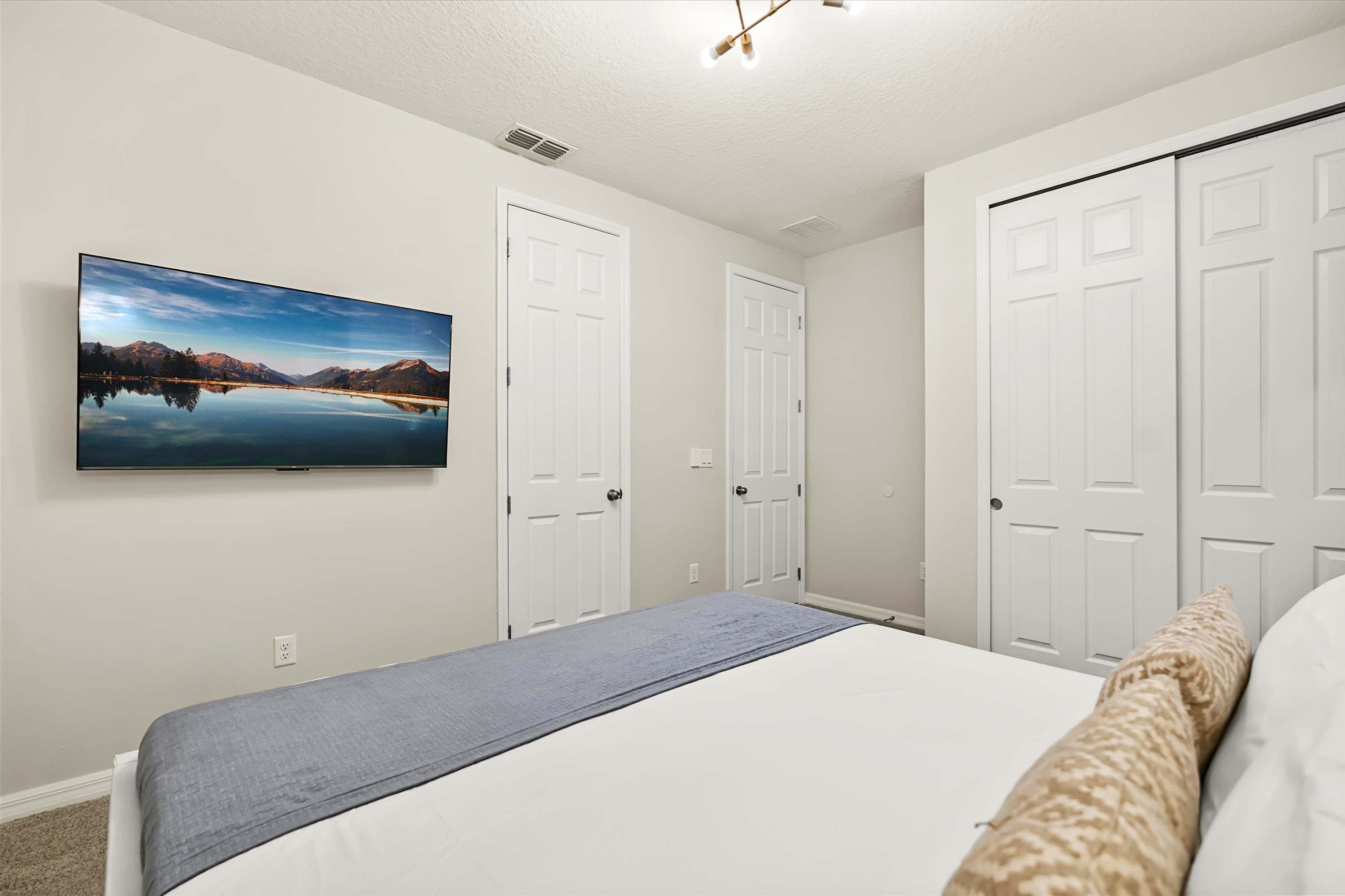 View of the modern bedroom showing a wall-mounted TV, two white doors, and a large closet with bi-fold doors, all against light-colored walls.