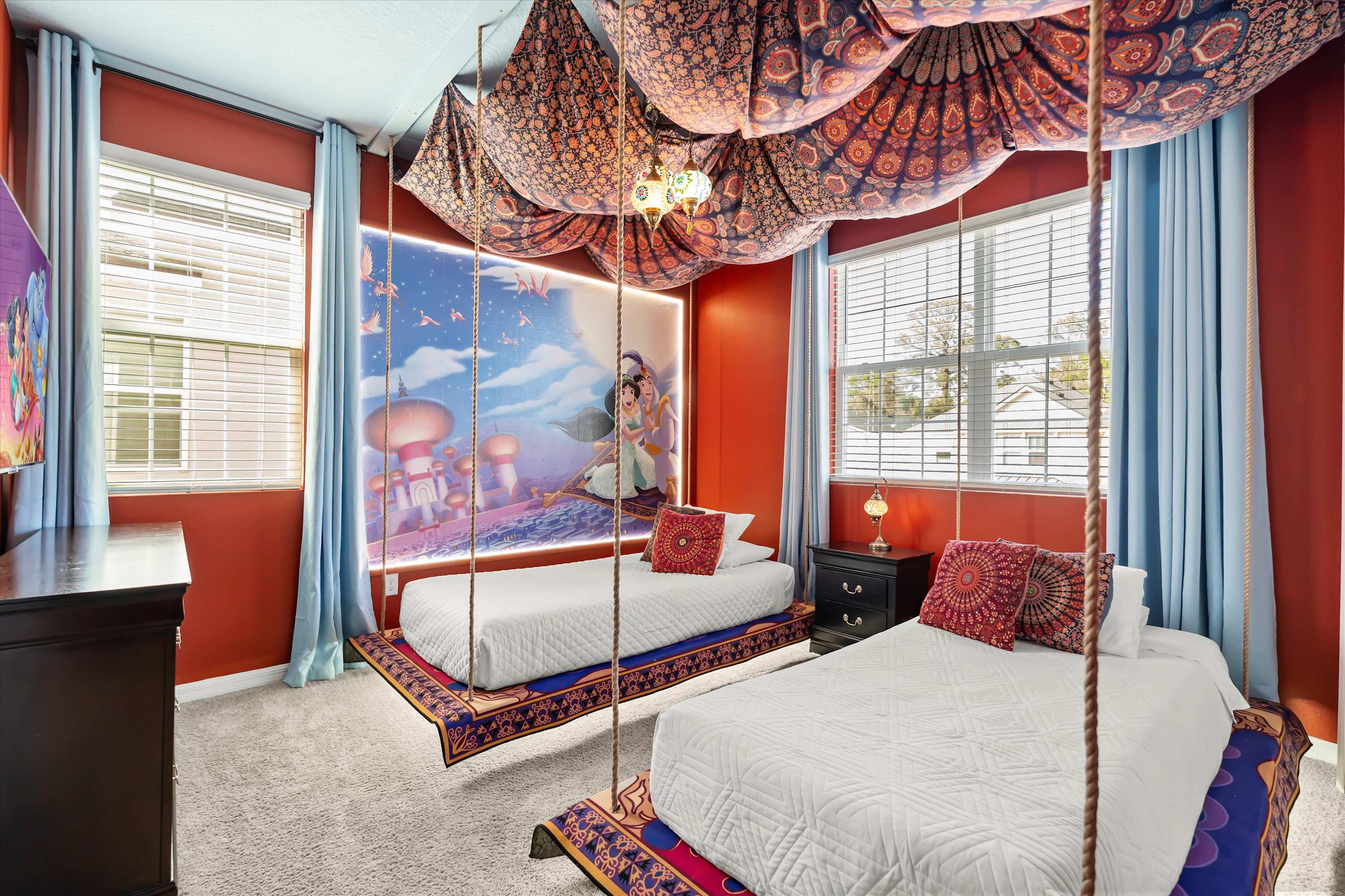 Vibrant Aladdin-themed bedroom with two hanging twin beds, red walls, blue curtains, and a large mural depicting Aladdin and Jasmine on a magic carpet.