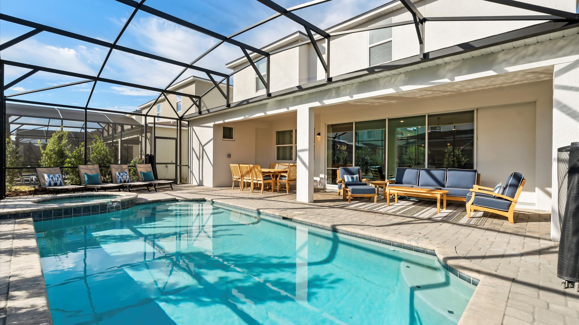 Screened-in outdoor pool area with a large rectangular pool, a covered patio featuring a wooden dining table and a seating area with blue cushions, all set against a modern house exterior.