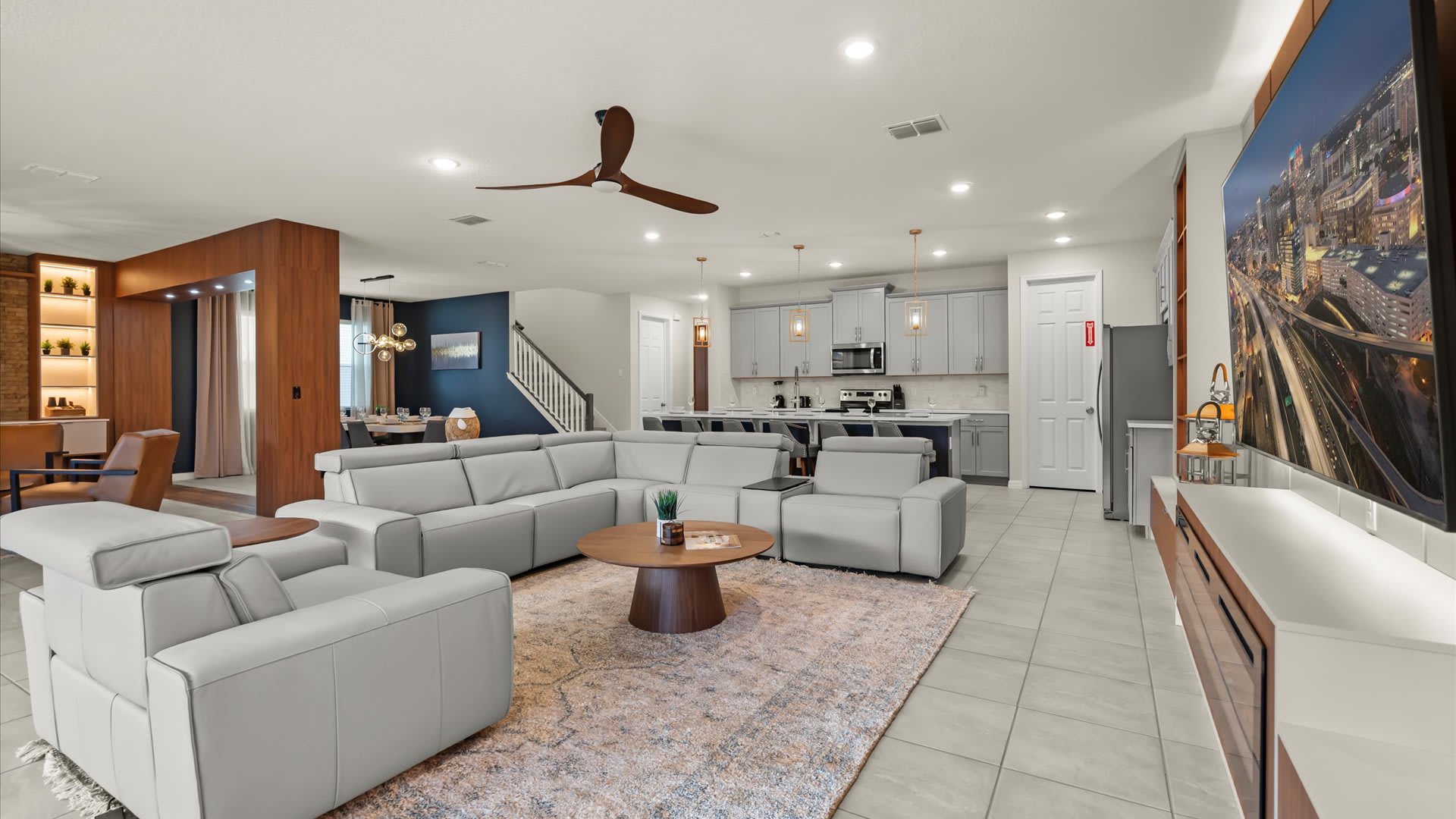 Spacious open-concept living room with a light gray sectional sofa, two armchairs, a round wooden coffee table, and a large wall-mounted TV above a modern fireplace, with a kitchen visible in the background.
