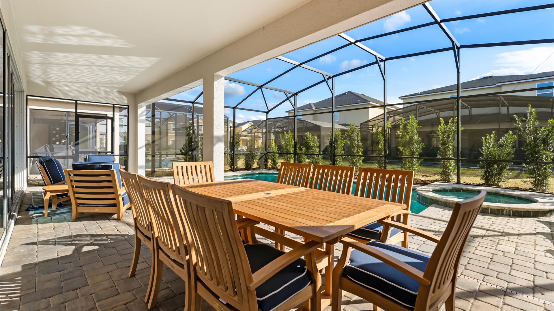 Covered outdoor patio with a large wooden dining table and chairs with blue cushions, overlooking a private screened pool area and backyard, with a modern house exterior visible through large glass doors.