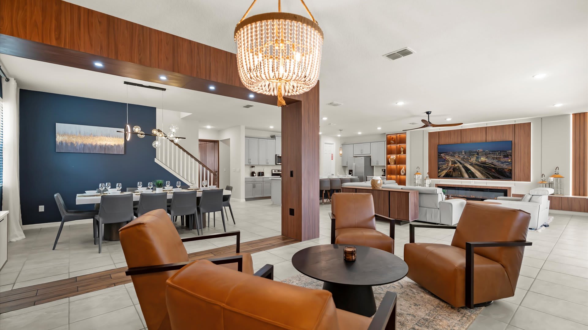 This wide shot captures an open-concept living space, showcasing a lounge area with leather chairs in the foreground, a dining area with a long table and blue accent wall, and a glimpse of the kitchen and living room beyond. Wood paneling frames the transitions between spaces, and a grand chandelier hangs above the lounge.