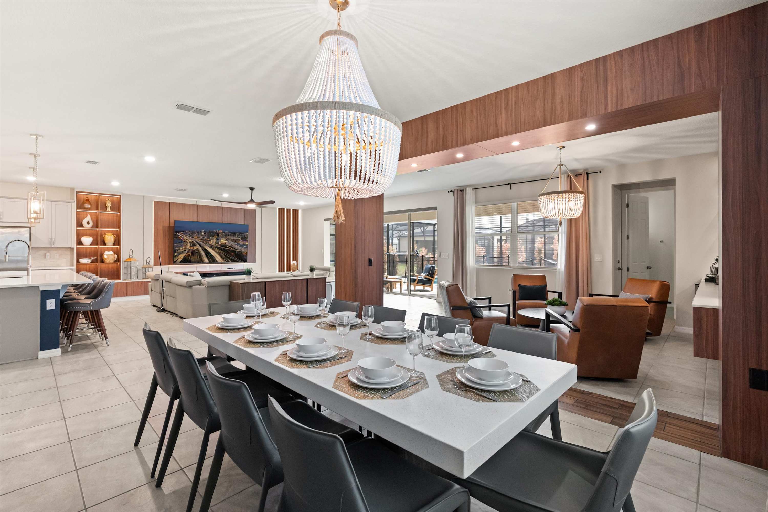 This wide view of the dining area shows a long white table set for eight, with a grand crystal chandelier overhead. The space opens to the kitchen on the left and the living room in the background, with wood paneling defining the architectural elements.