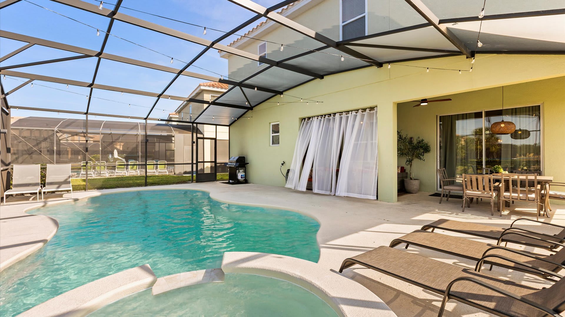 Large screened pool deck with a sparkling blue pool and hot tub, adjacent to a covered patio area furnished with an outdoor dining set and a BBQ grill, offering direct access to the house.