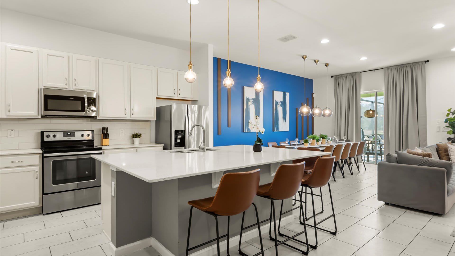 A wide shot of the modern kitchen with white shaker cabinets and stainless steel appliances, featuring a large island with bar seating. The kitchen seamlessly connects to the dining area with its vibrant blue accent wall, creating an open and inviting space.