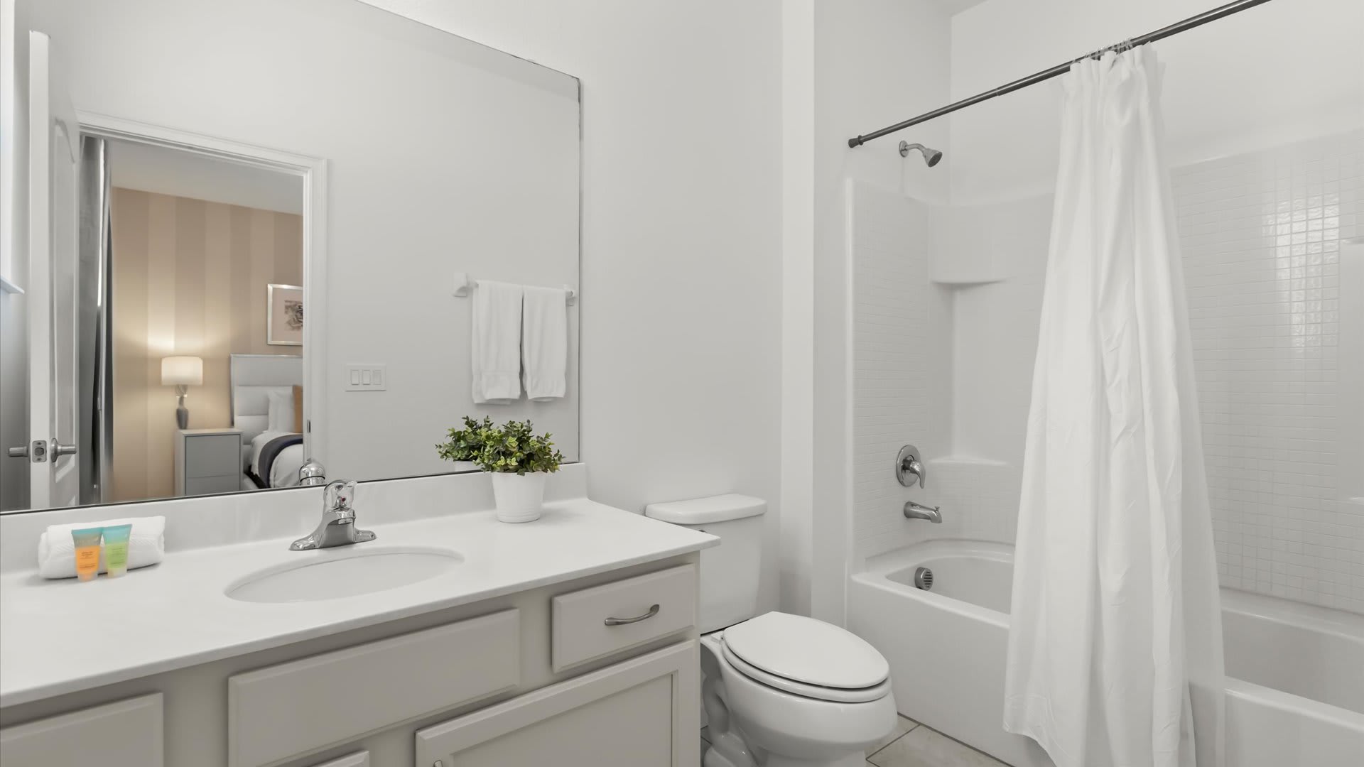 A clean, functional bathroom with a single vanity, white countertop, light gray cabinets, and a toilet. A shower-tub combination with a white curtain is visible, and a small potted plant decorates the counter.