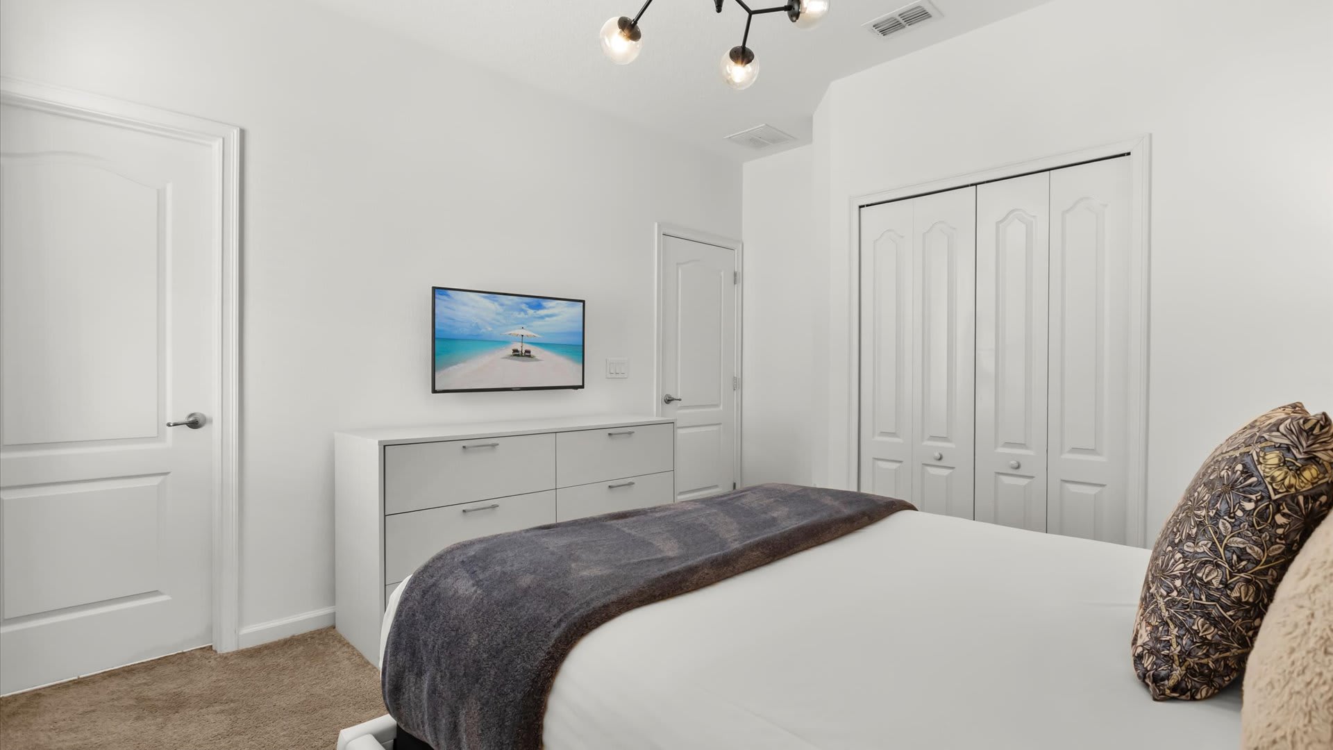 A modern bedroom with a white dresser and a wall-mounted flat-screen TV displaying a beach scene. The room features light-colored walls, a carpeted floor, and a closet with bi-fold doors.