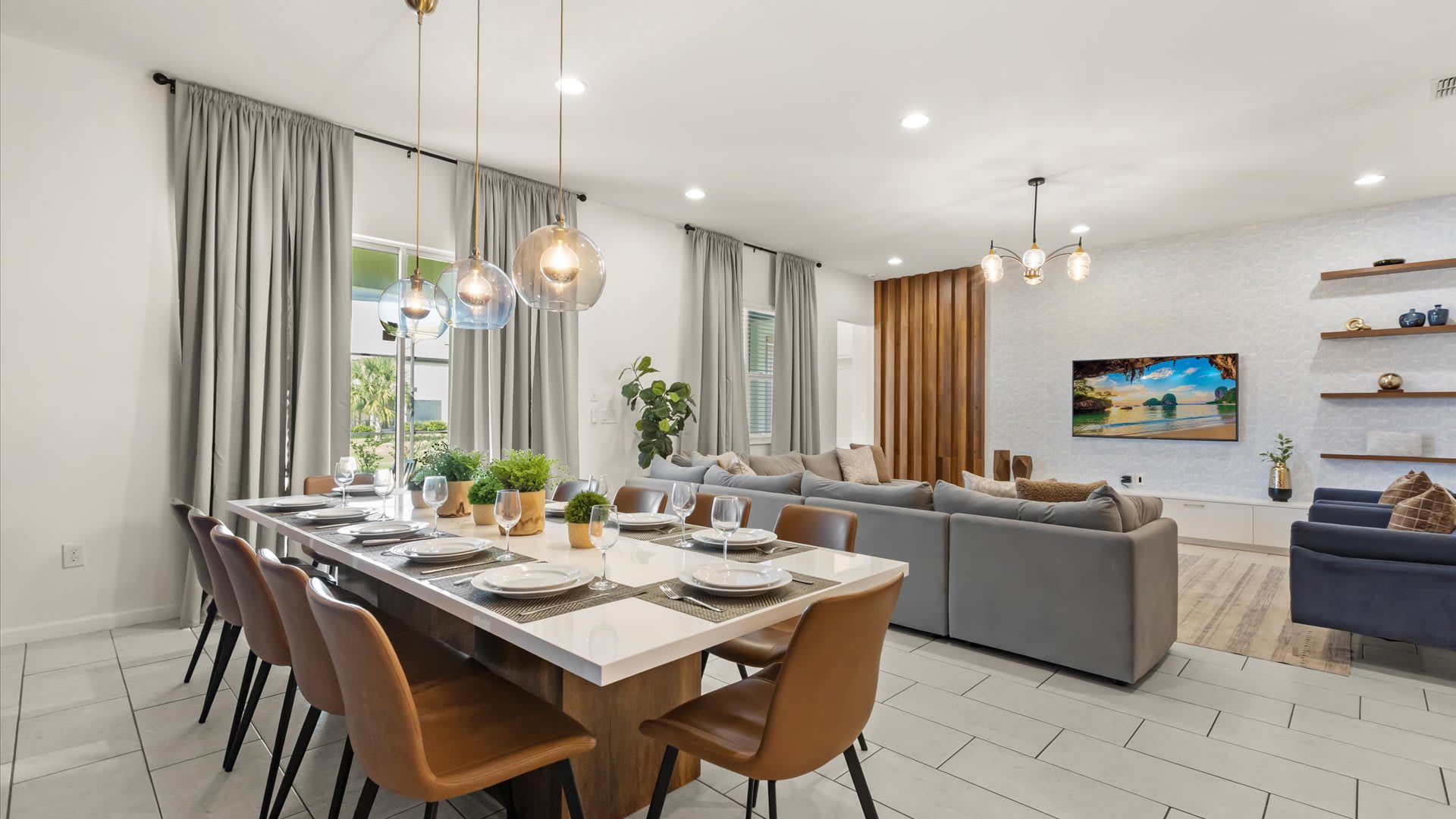 Spacious open-concept dining area with a long white table seating ten, illuminated by modern glass pendant lights, flowing into a bright living room with a large gray sectional sofa and a wall-mounted TV.