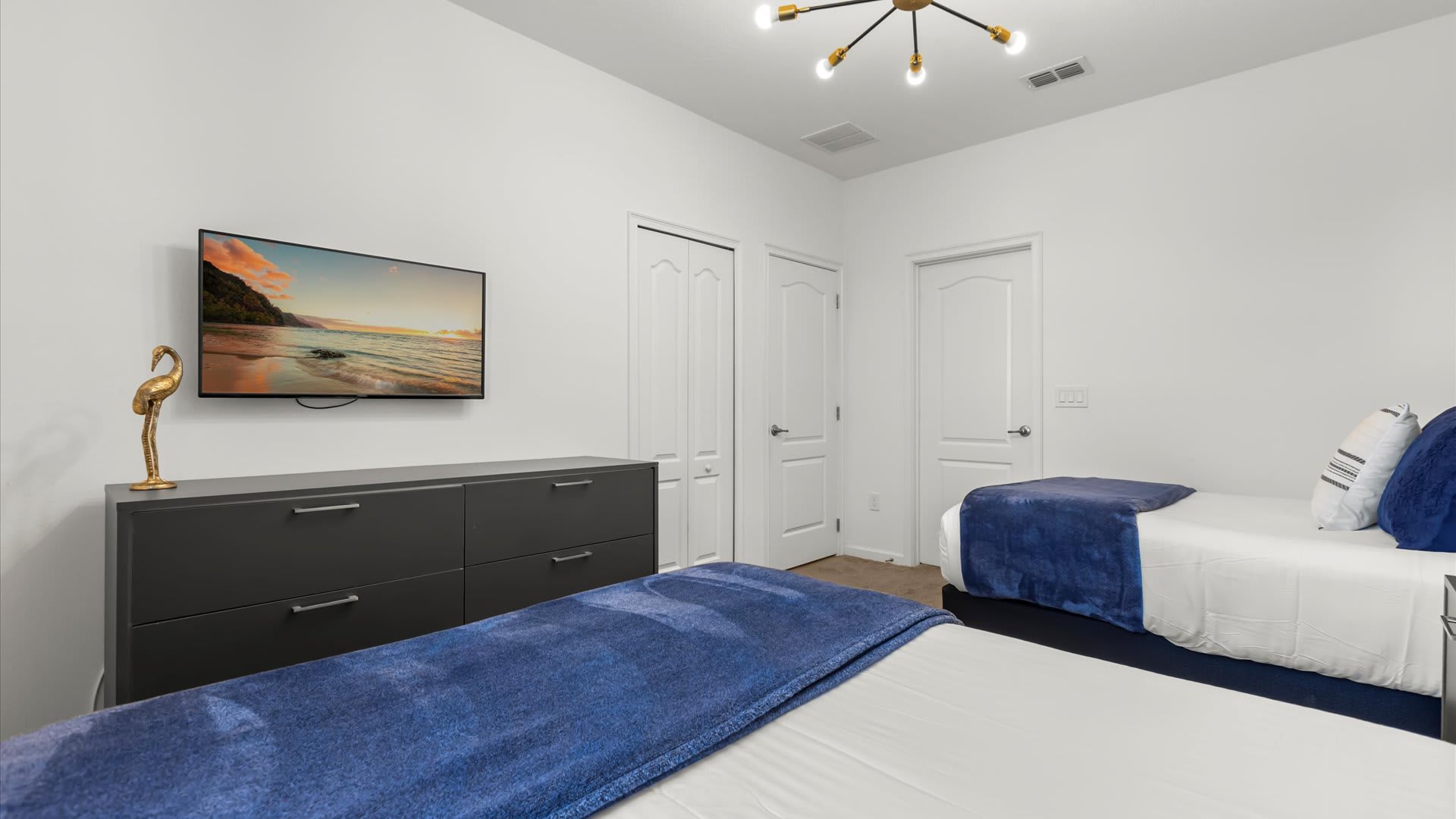 View of the bedroom with two twin beds, showcasing a wall-mounted TV above a gray dresser, and white bi-fold closet doors.