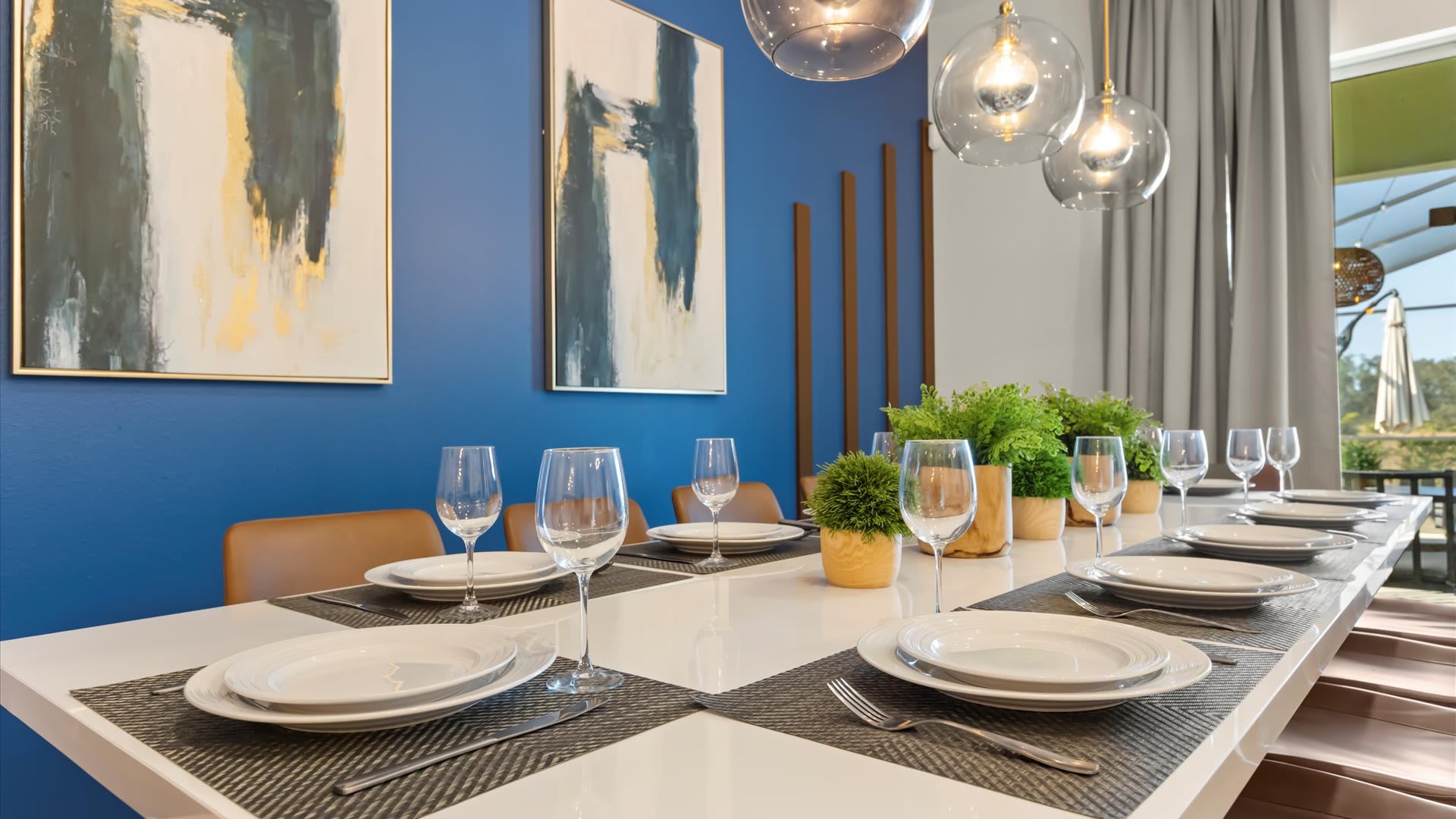 A close-up of the modern dining table set for ten, featuring white plates, wine glasses, and green plant centerpieces. The vibrant blue accent wall with abstract art and wooden slats provides a stylish backdrop, illuminated by three contemporary pendant lights.