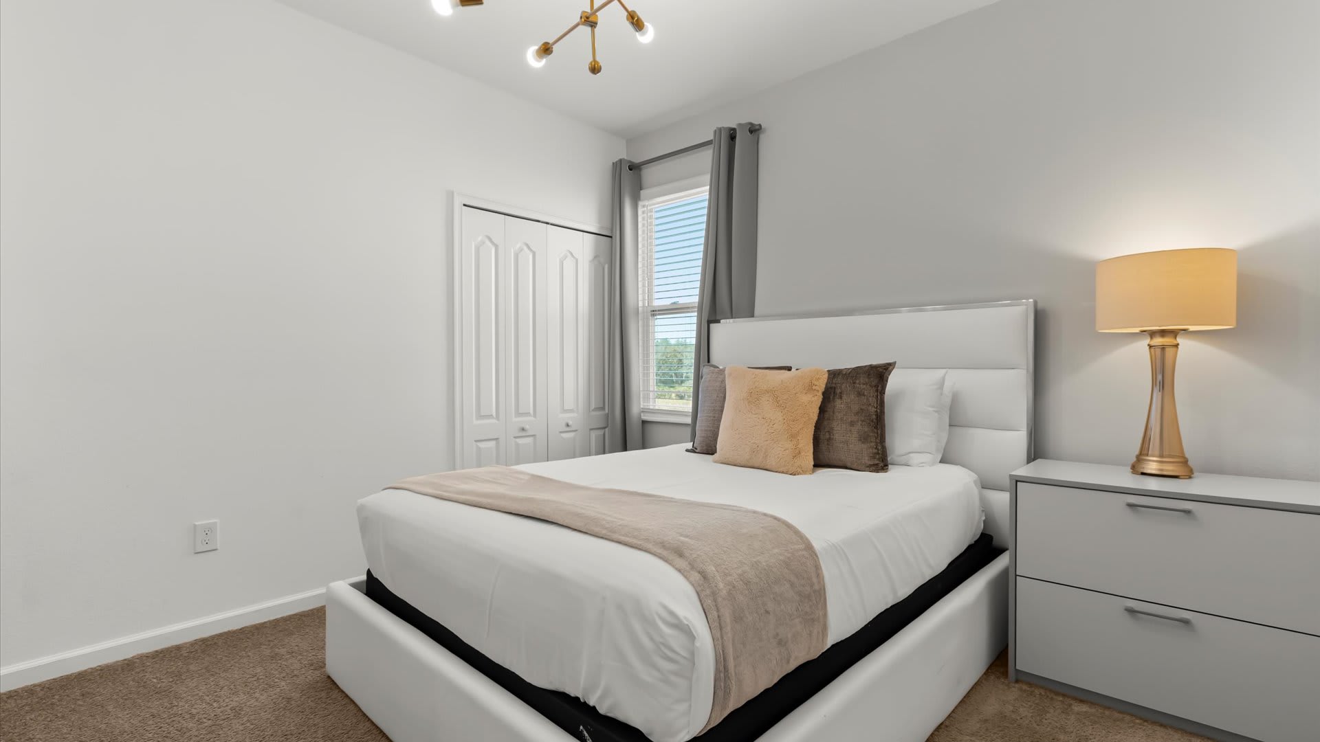 A bright bedroom featuring a queen-sized bed with a white upholstered headboard, a gray nightstand with a gold-based lamp, and a window with gray curtains. The room has light walls and a carpeted floor.