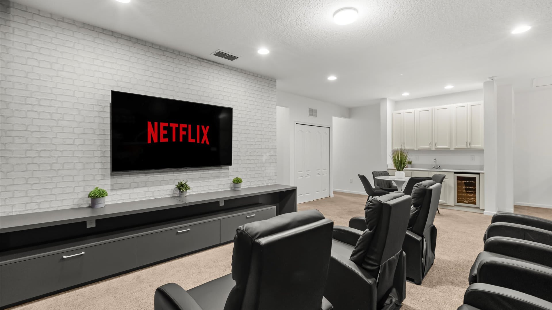 A modern media room features black leather recliners facing a large wall-mounted TV displaying "NETFLIX," set against a white brick accent wall. A wet bar with white cabinets, a sink, and a beverage cooler is visible in the background, complementing the neutral carpeted floor.