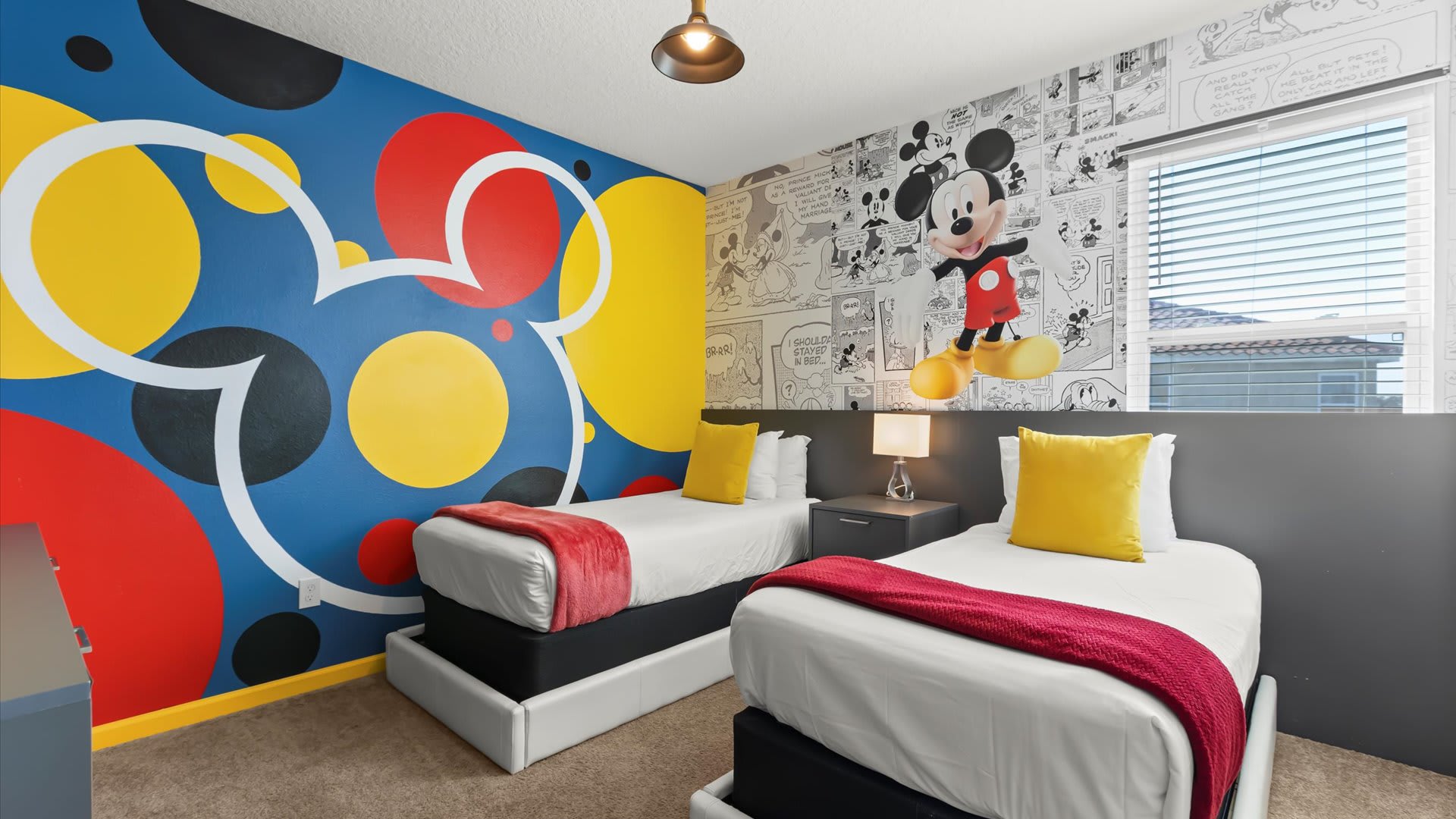 Vibrant Mickey Mouse themed bedroom featuring two twin beds, a wall with large red, yellow, and blue Mickey head silhouettes, and another wall with comic strip wallpaper and a 3D Mickey figure.