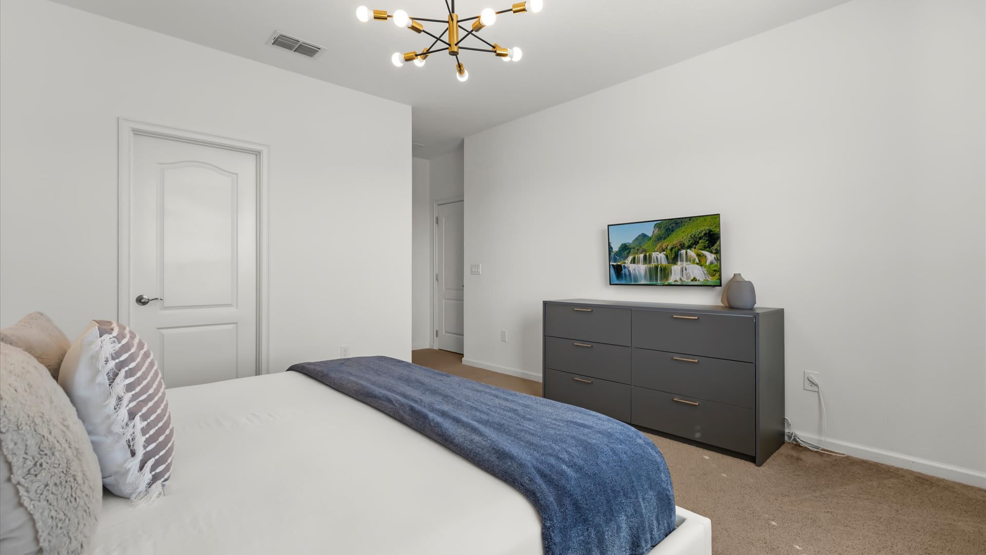 View of the foot of the bed in bedroom-1, showing a dark gray dresser with a wall-mounted flat-screen TV displaying a waterfall scene. The room has white walls and a light brown carpet.