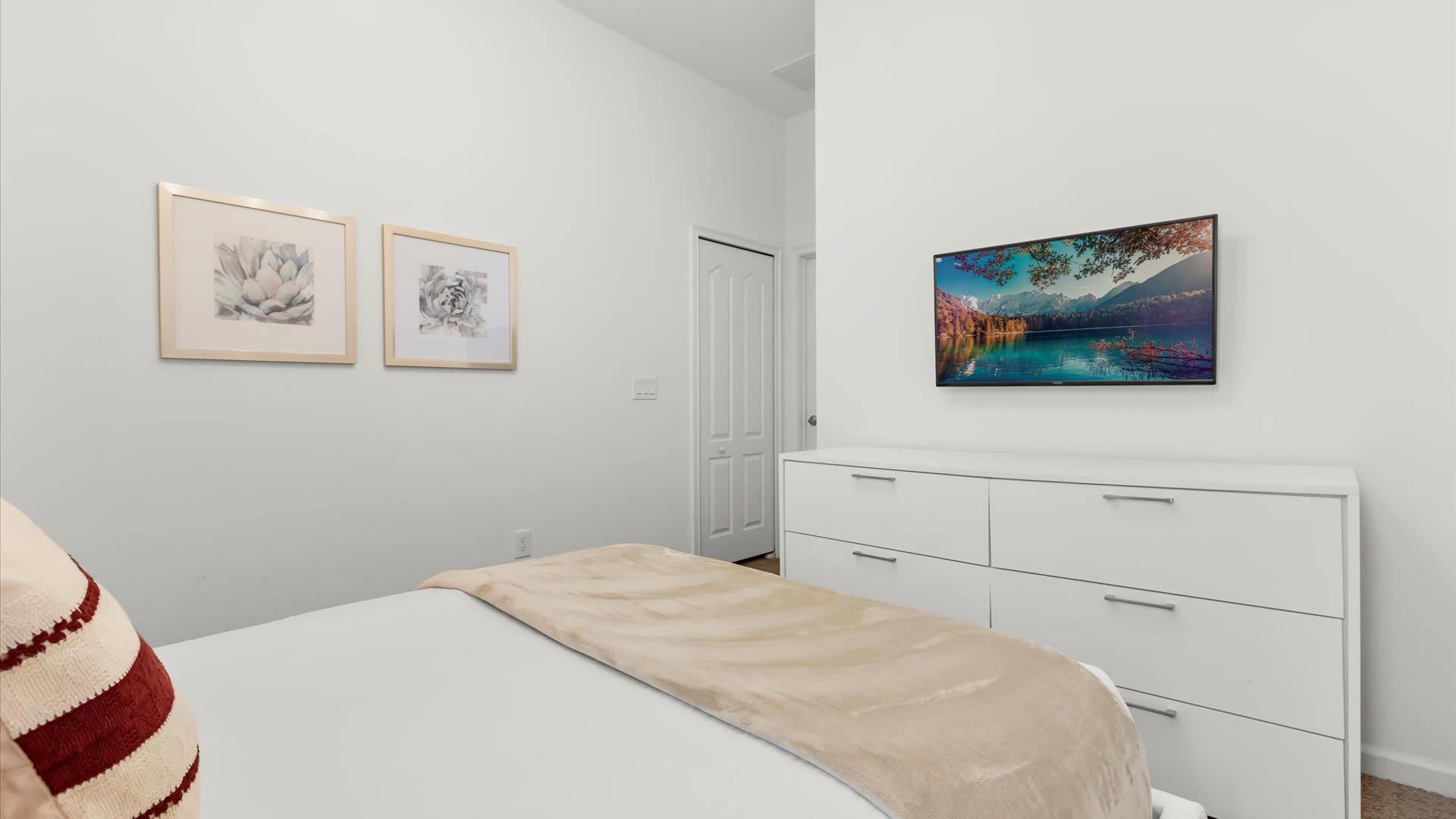 A clean bedroom featuring a flat-screen TV mounted above a white dresser with multiple drawers, positioned opposite a bed with a beige throw blanket. Two framed artworks hang on the wall.