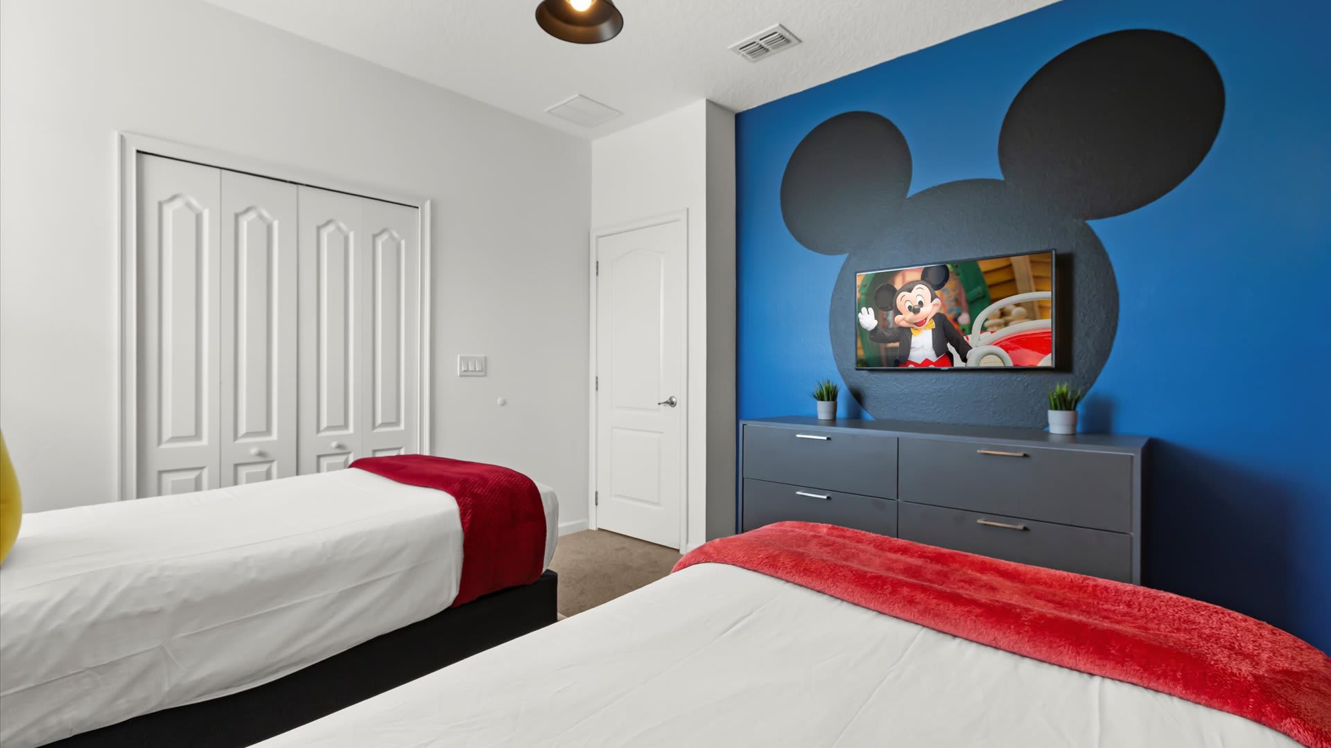 View of the Mickey Mouse themed bedroom showing a wall-mounted TV against a blue wall with a large Mickey head silhouette, a gray dresser, and white bi-fold closet doors.