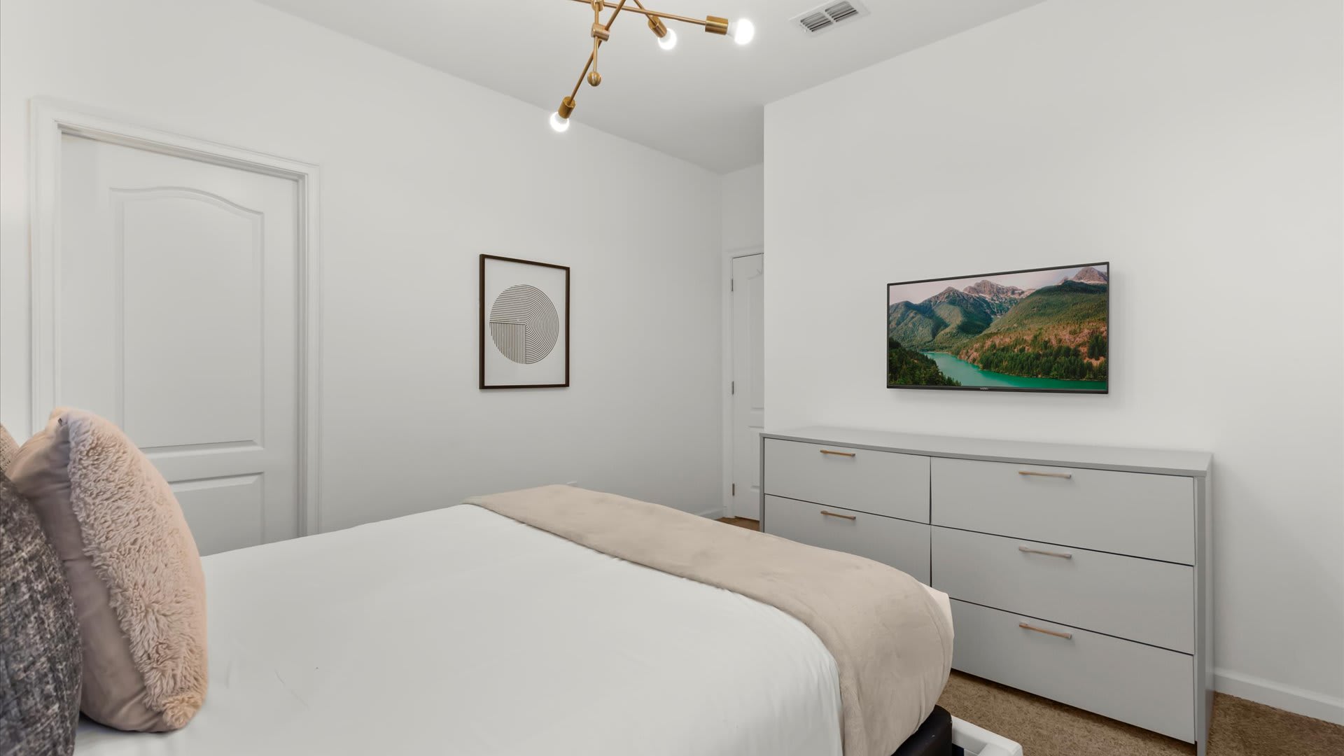 A minimalist bedroom featuring a white dresser with gold pulls and a wall-mounted flat-screen TV displaying a scenic mountain lake. The room has light walls, a carpeted floor, and a modern light fixture.